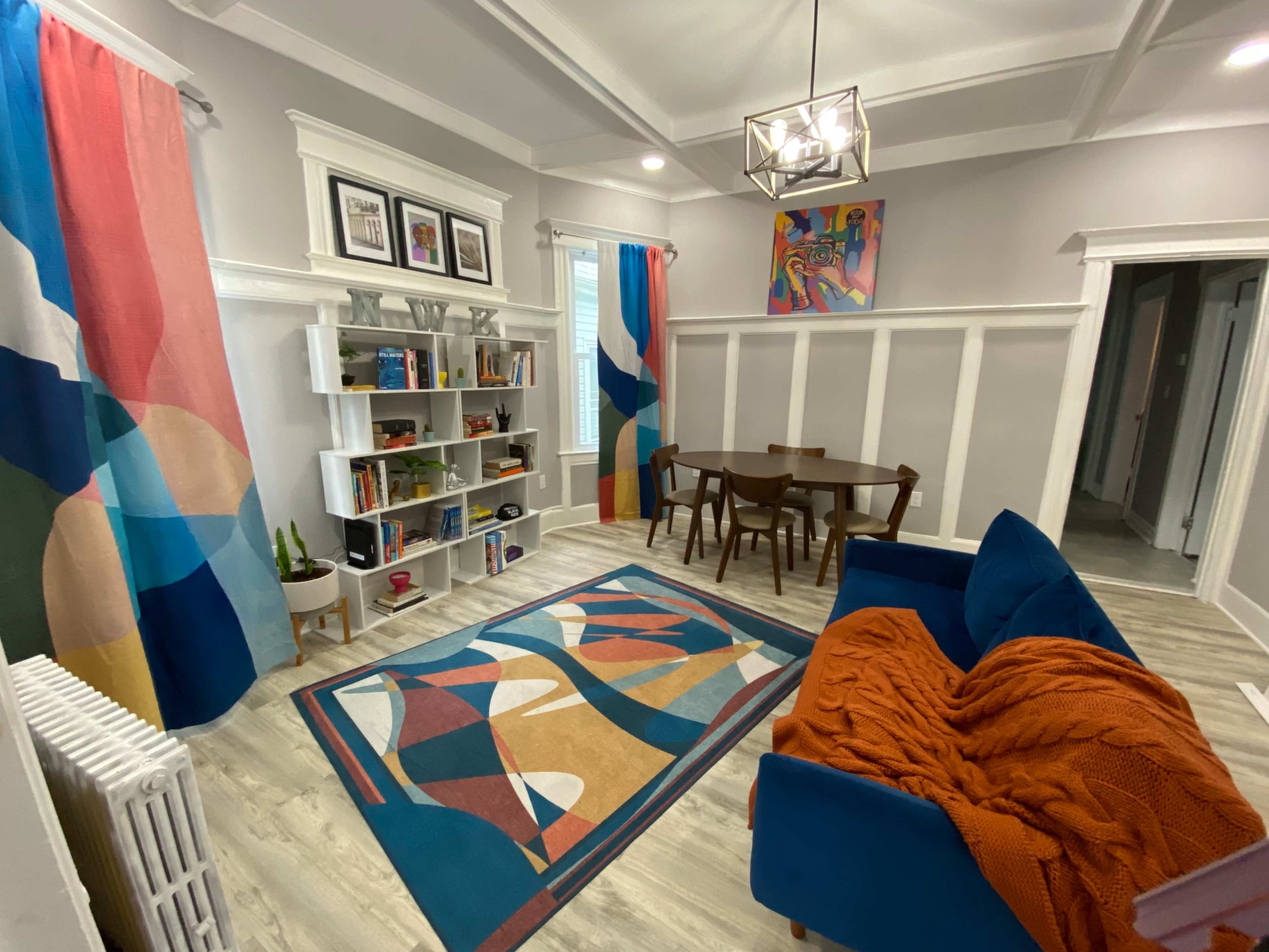 The image shows a modern living space featuring a blue couch, a patterned rug, a wooden dining table with chairs, and colorful curtains.