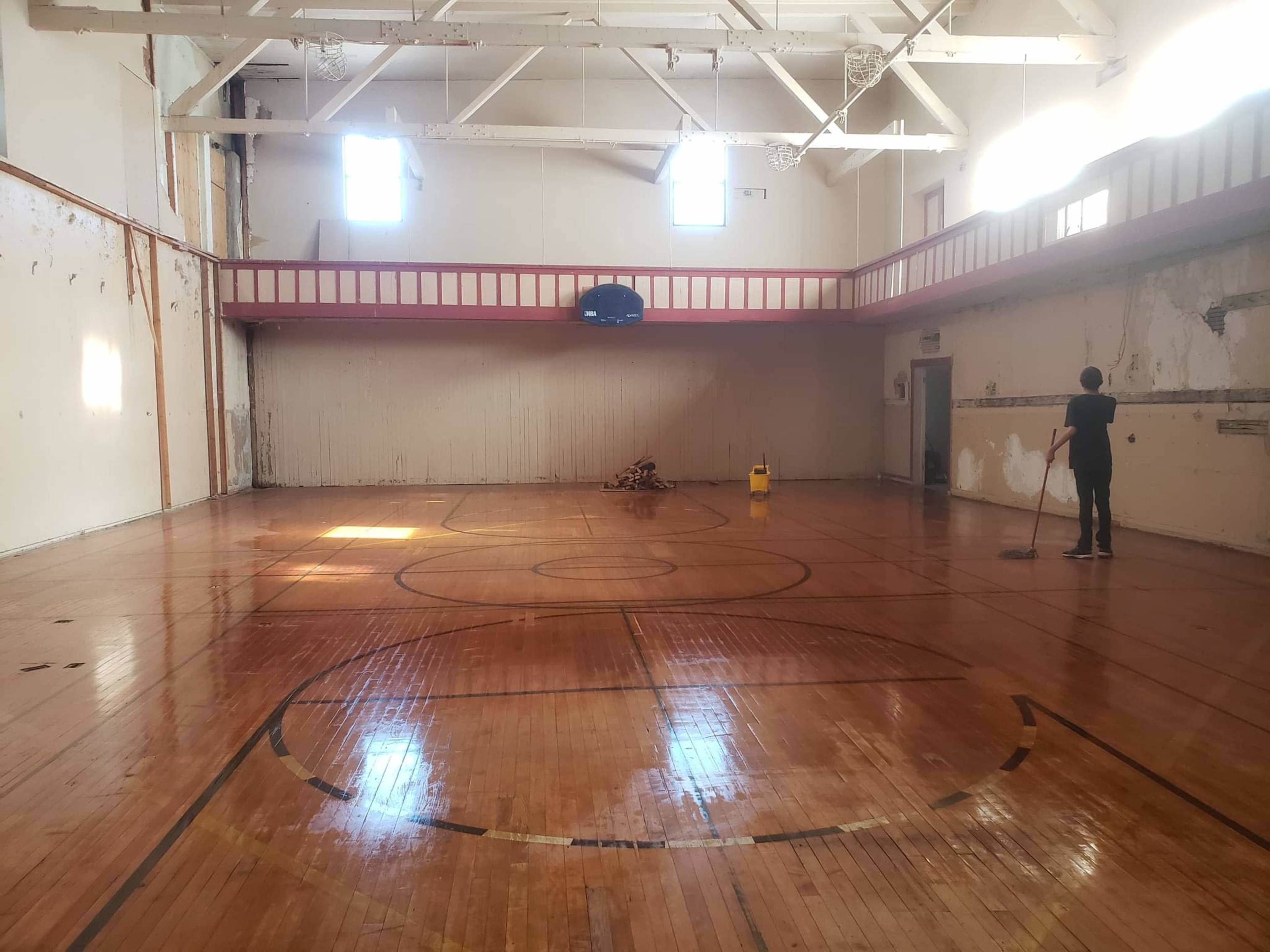 A person is mopping the wooden floor of a vacant gymnasium with exposed walls and a basketball hoop in the background.