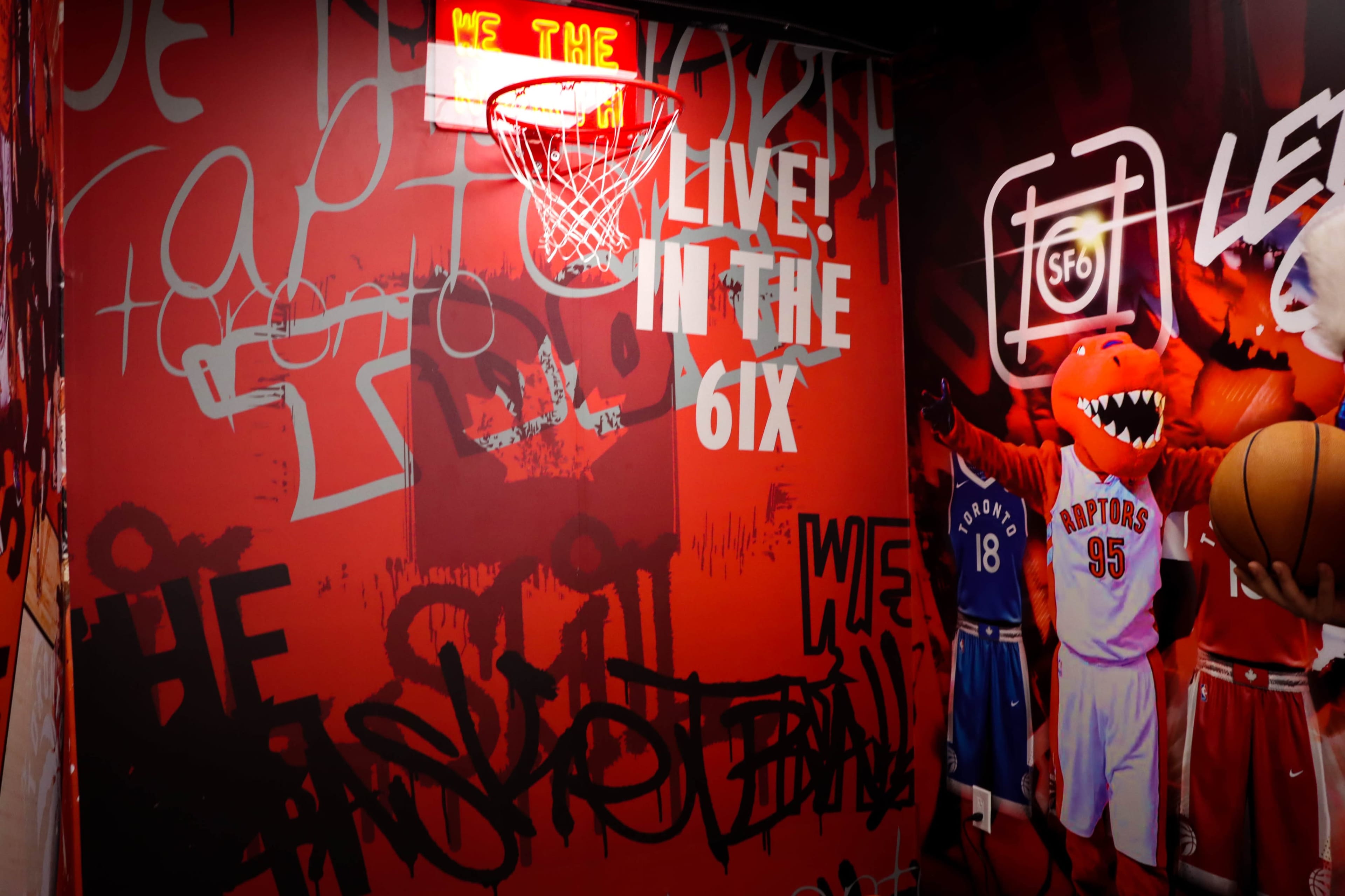 The image features a basketball hoop surrounded by vibrant murals and graffiti in a room decorated with Toronto Raptors themes.