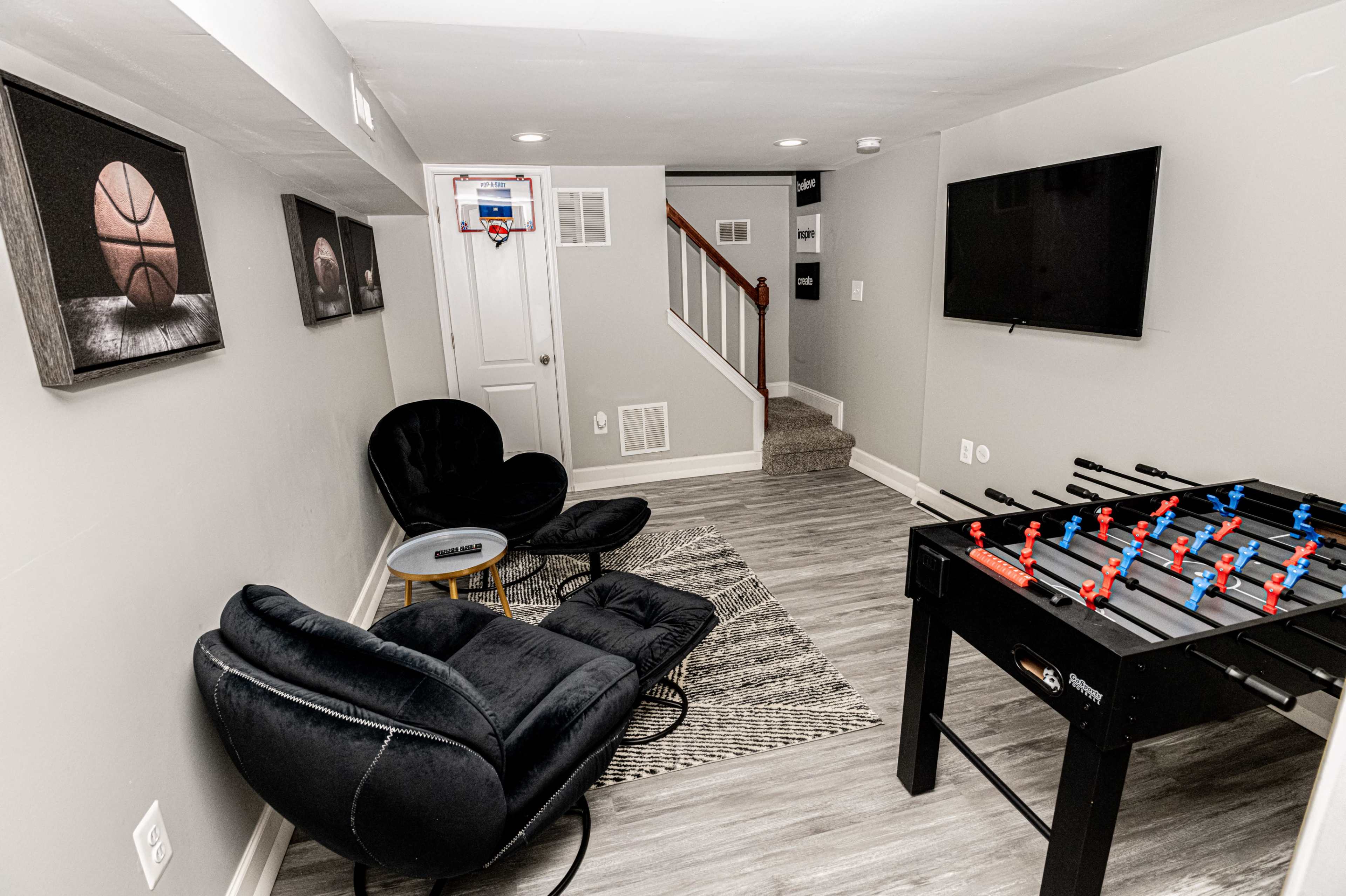 The image shows a basement recreation area featuring a foosball table, two black chairs, a patterned rug, and a small wall-mounted basketball hoop.