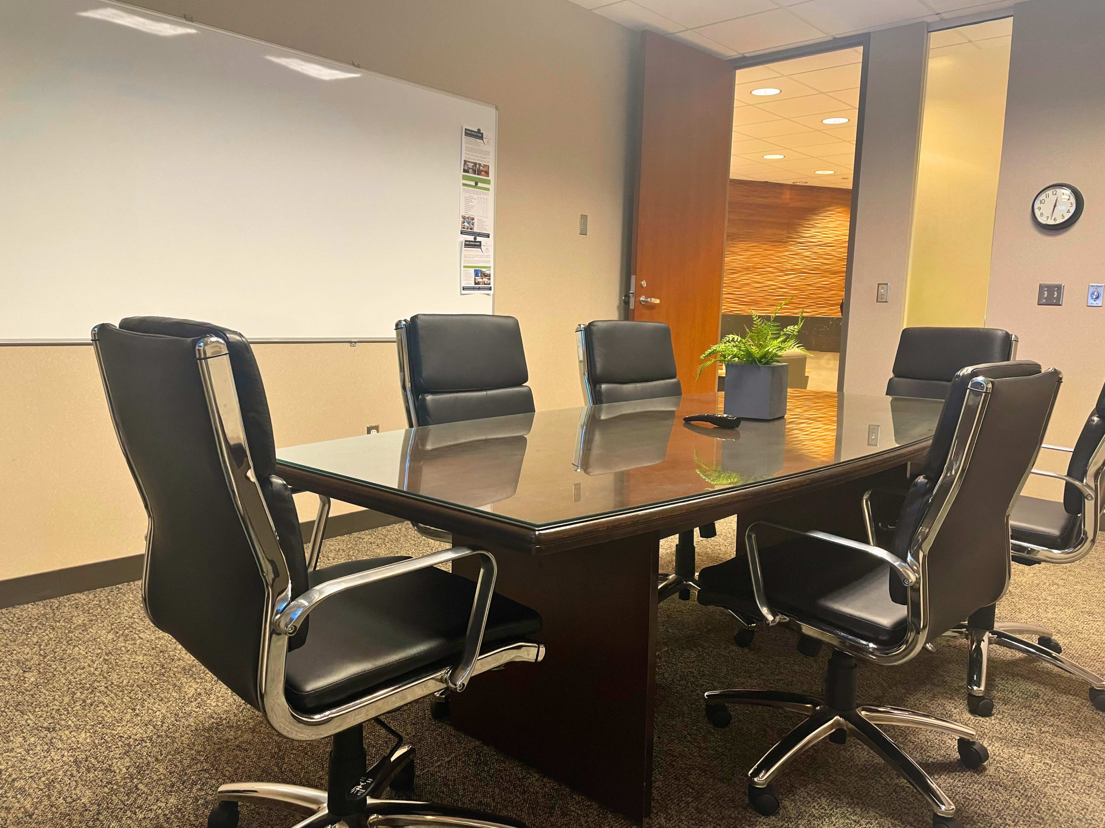 A conference room featuring a large rectangular table surrounded by black leather chairs, with a whiteboard and a plant in the center.