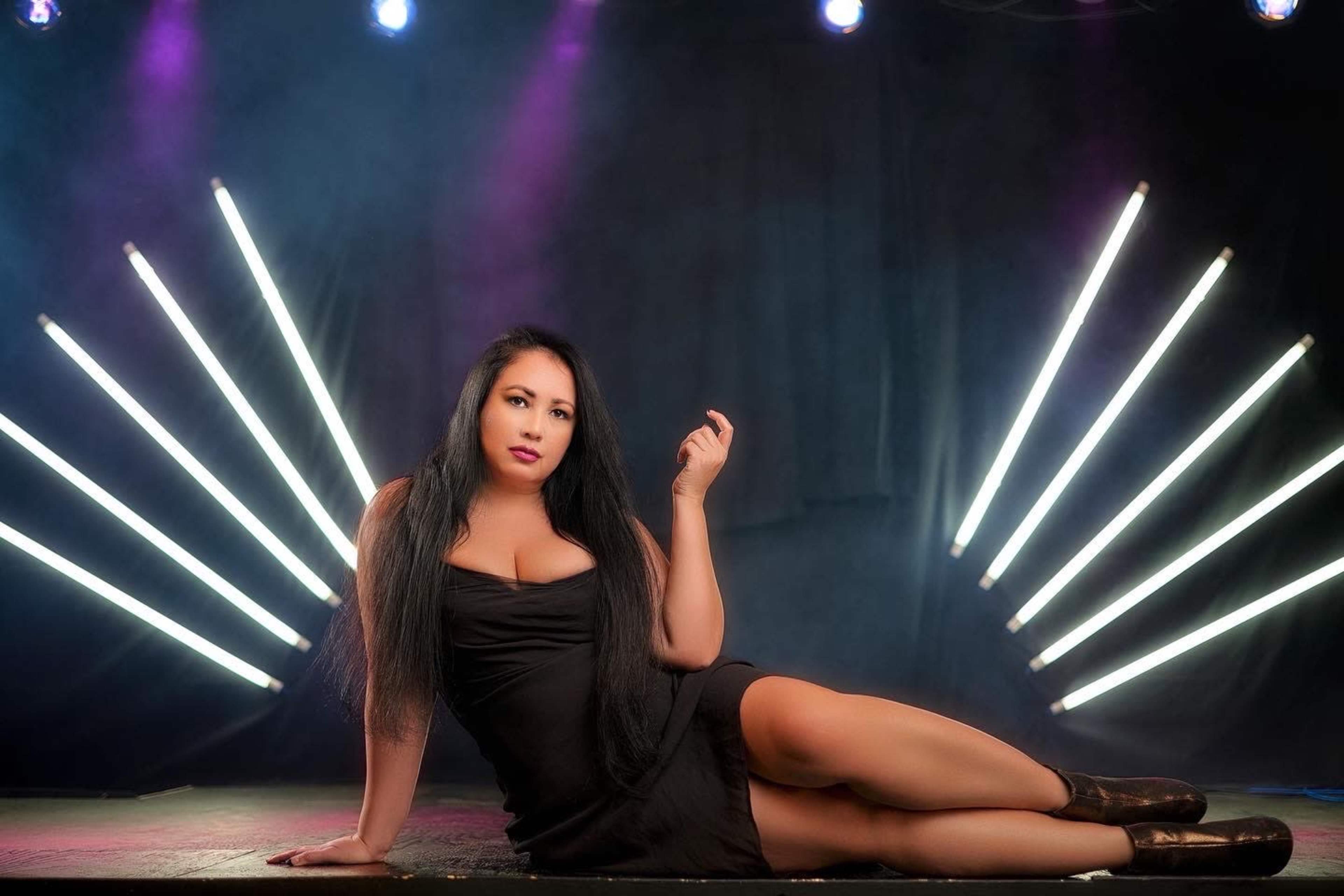 A woman in a black dress lounges on a stage with dramatic lighting from angled neon strips in the background.