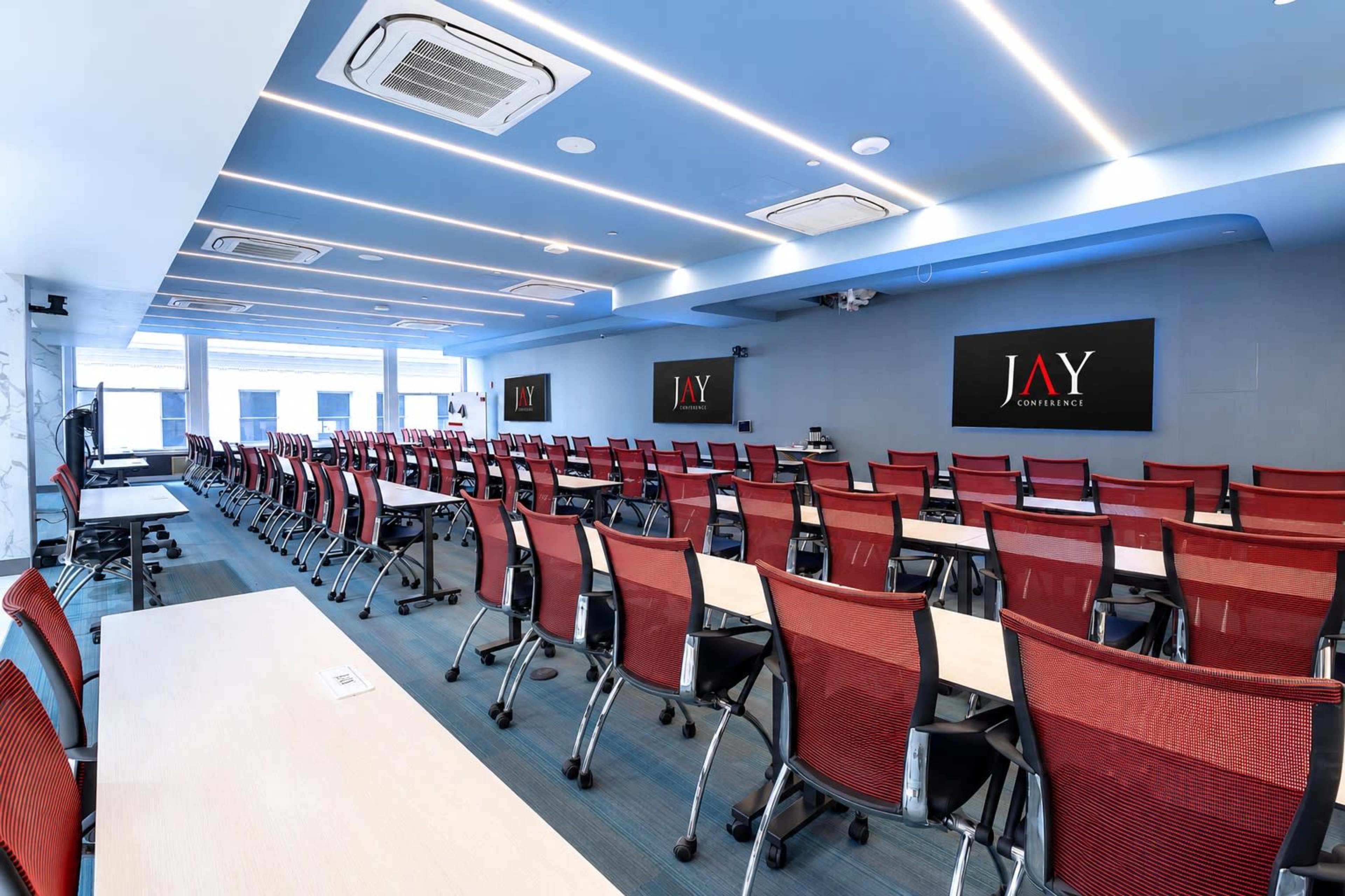 The image shows a spacious conference room with rows of red and black chairs, arranged facing a front area featuring two large screens displaying the text "JAY CONSULTING."