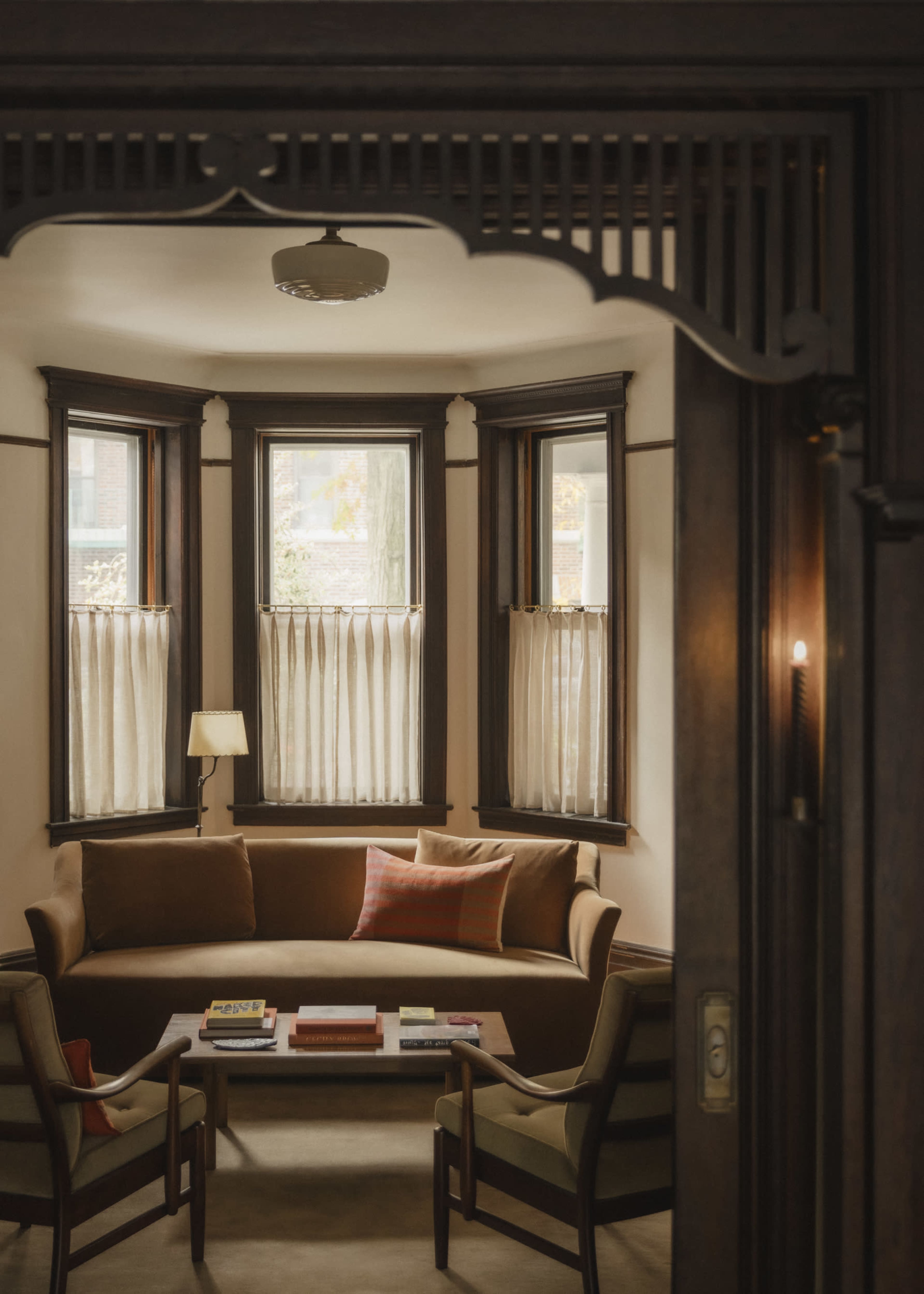 A cozy living room features a brown couch, two armchairs, and a coffee table, all framed by large windows with sheer curtains.