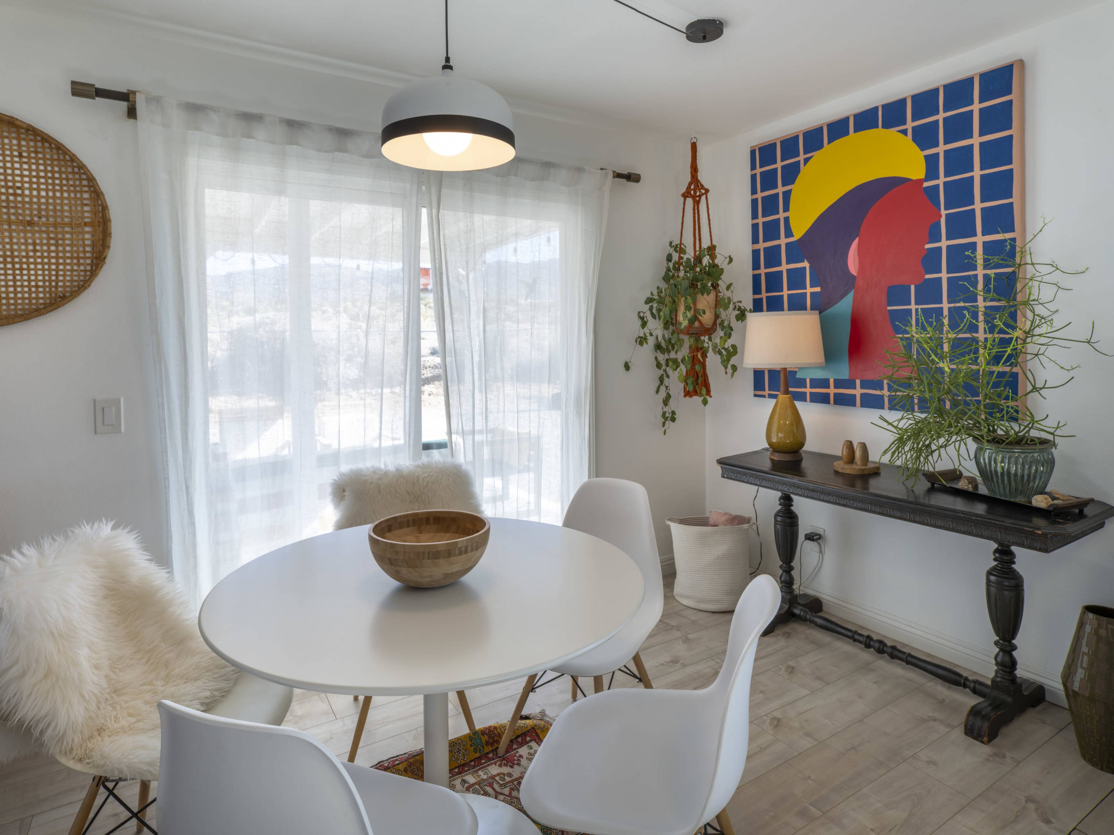 A modern dining area features a round white table surrounded by four chairs, with a large artwork on the wall and a window allowing natural light to enter.