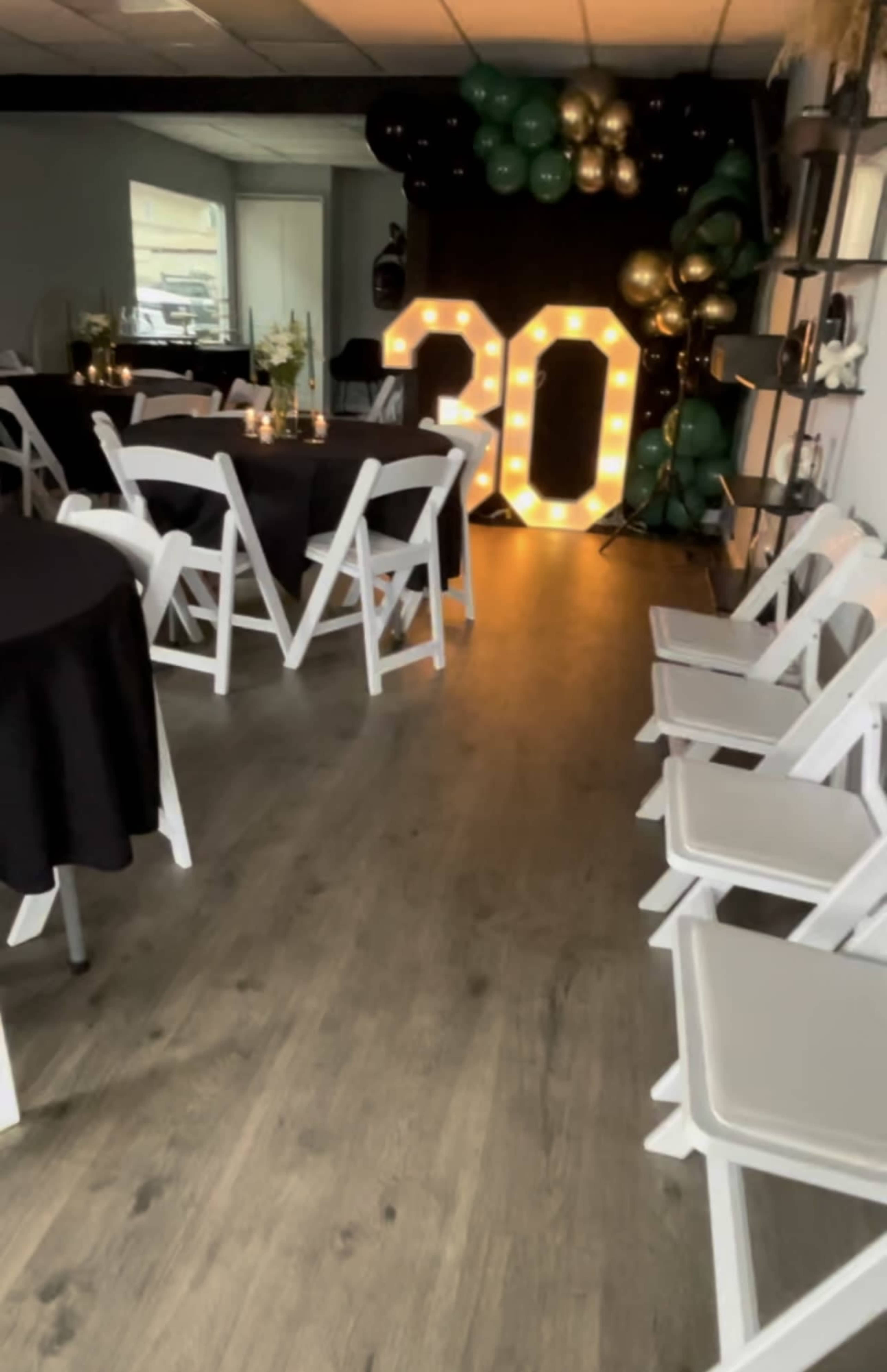 The image shows a decorated indoor party space featuring a large illuminated "30" sign, black and green balloons, and tables with black tablecloths arranged around the room.