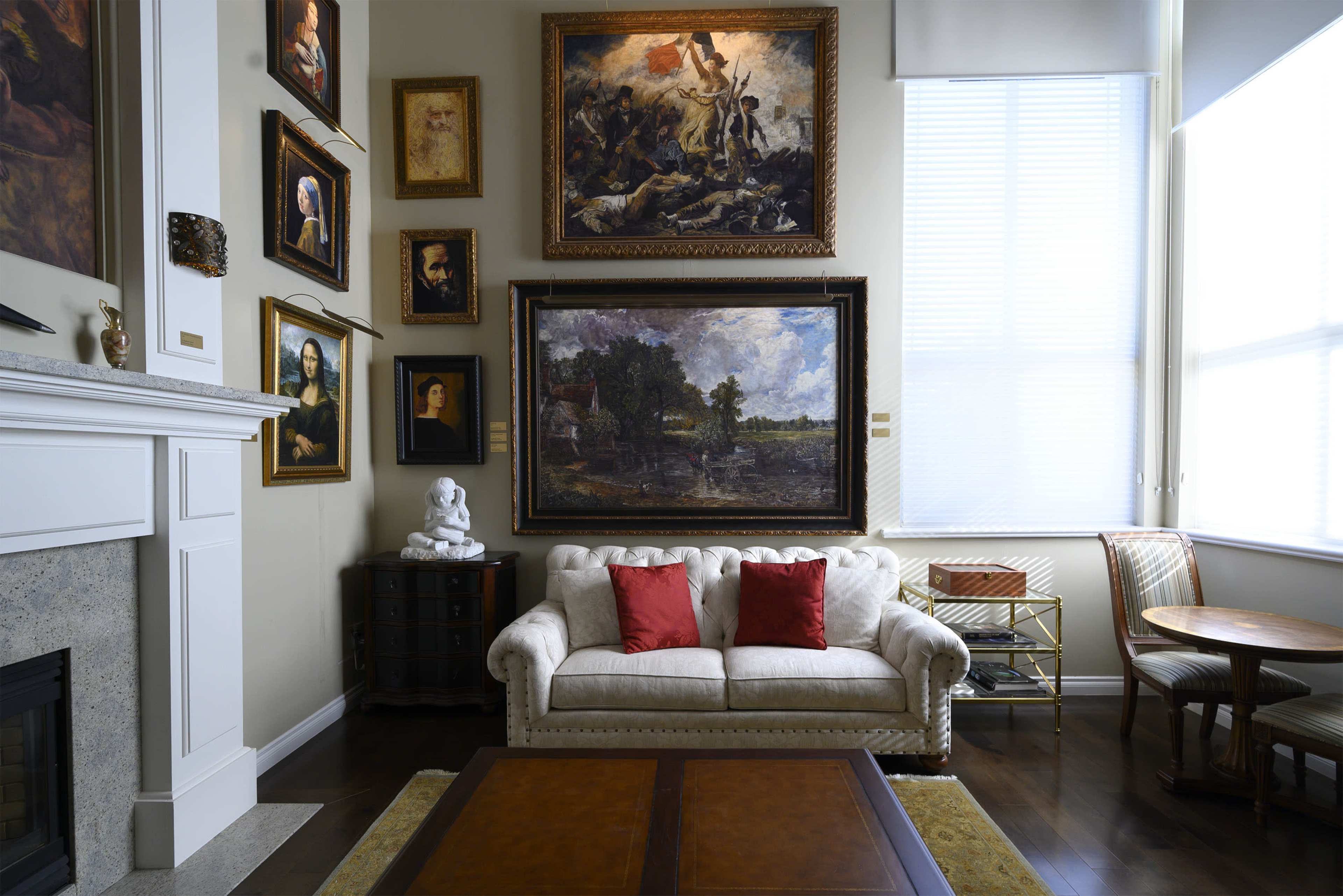 A living room with a white velvet sofa adorned with two red cushions, surrounded by various framed paintings and a statue, with large windows allowing natural light to enter.