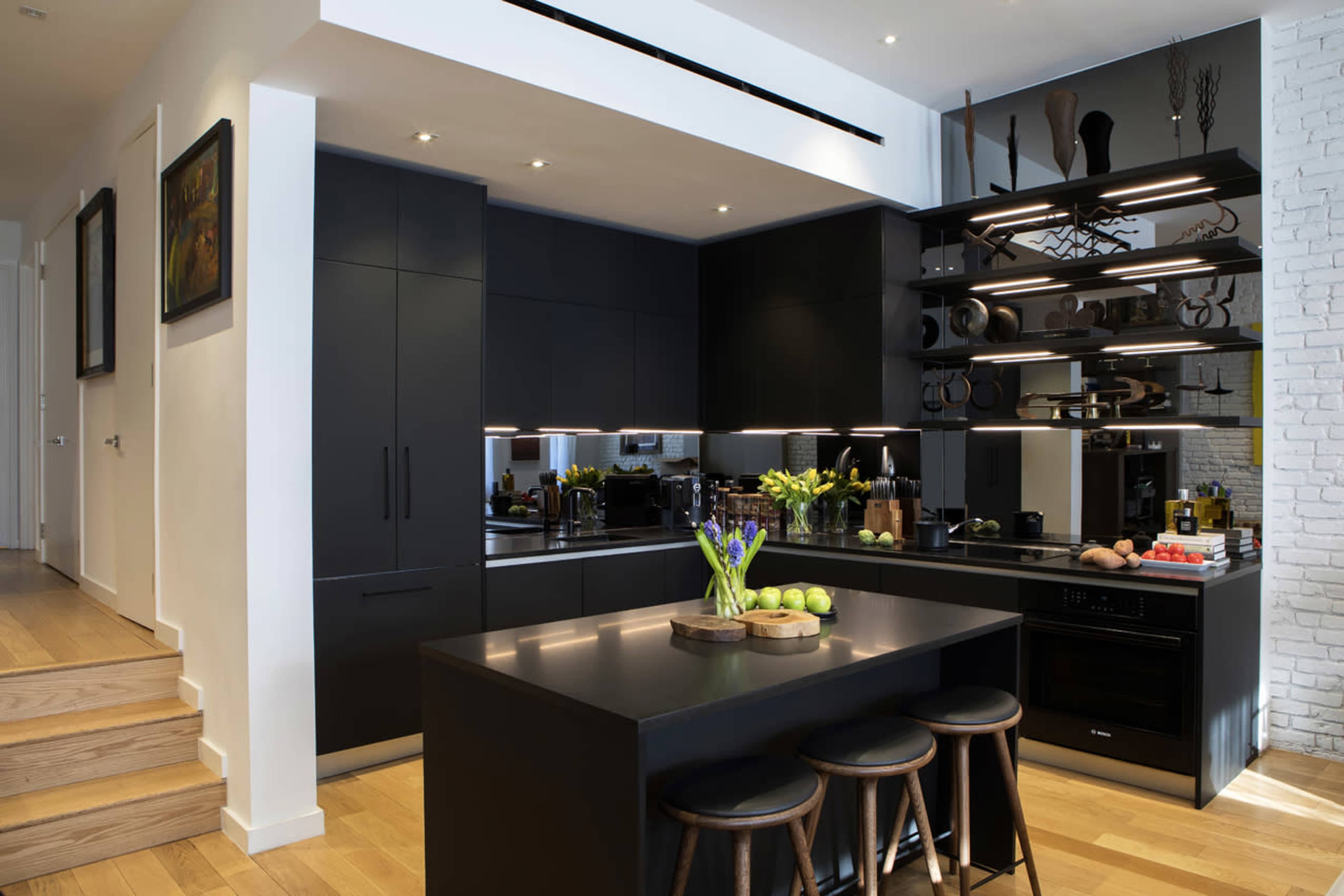 The image shows a modern kitchen with black cabinetry, a large island with three wooden stools, and open shelving displaying kitchenware against a white brick wall.