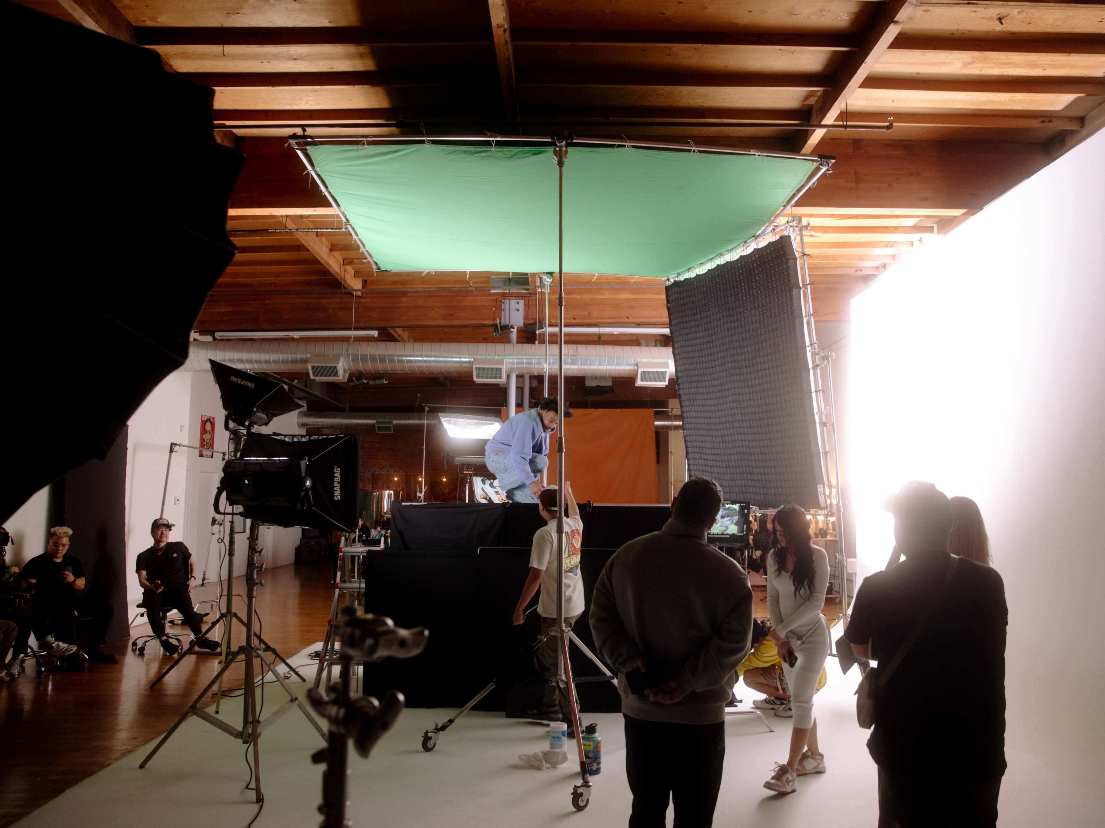 A film crew works on a studio set with lighting equipment and backdrop, while several people observe from the foreground.