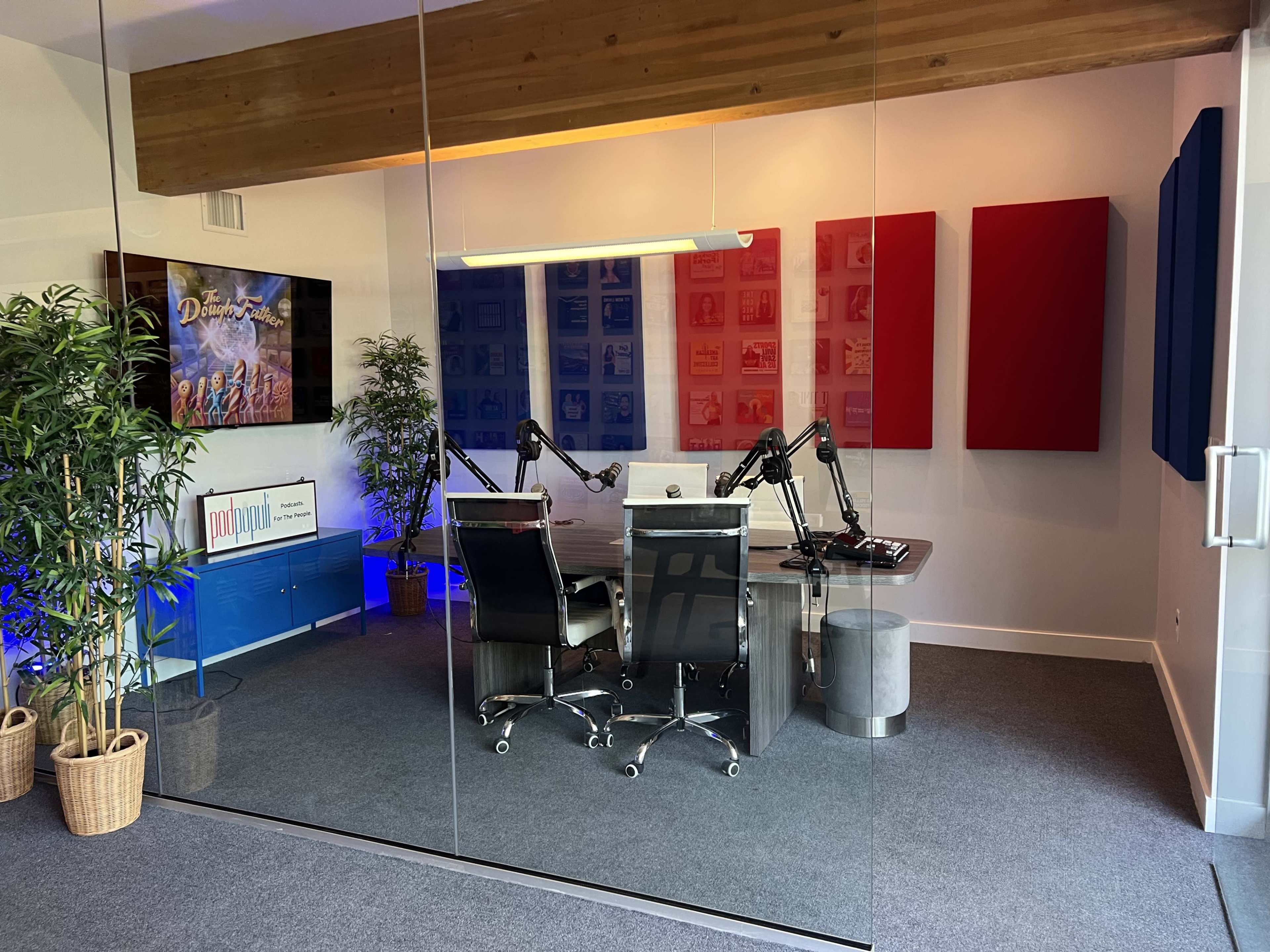 The image shows a modern podcasting studio featuring a glass wall, a large table with a computer, adjustable microphones, and colorful acoustic panels on the walls.