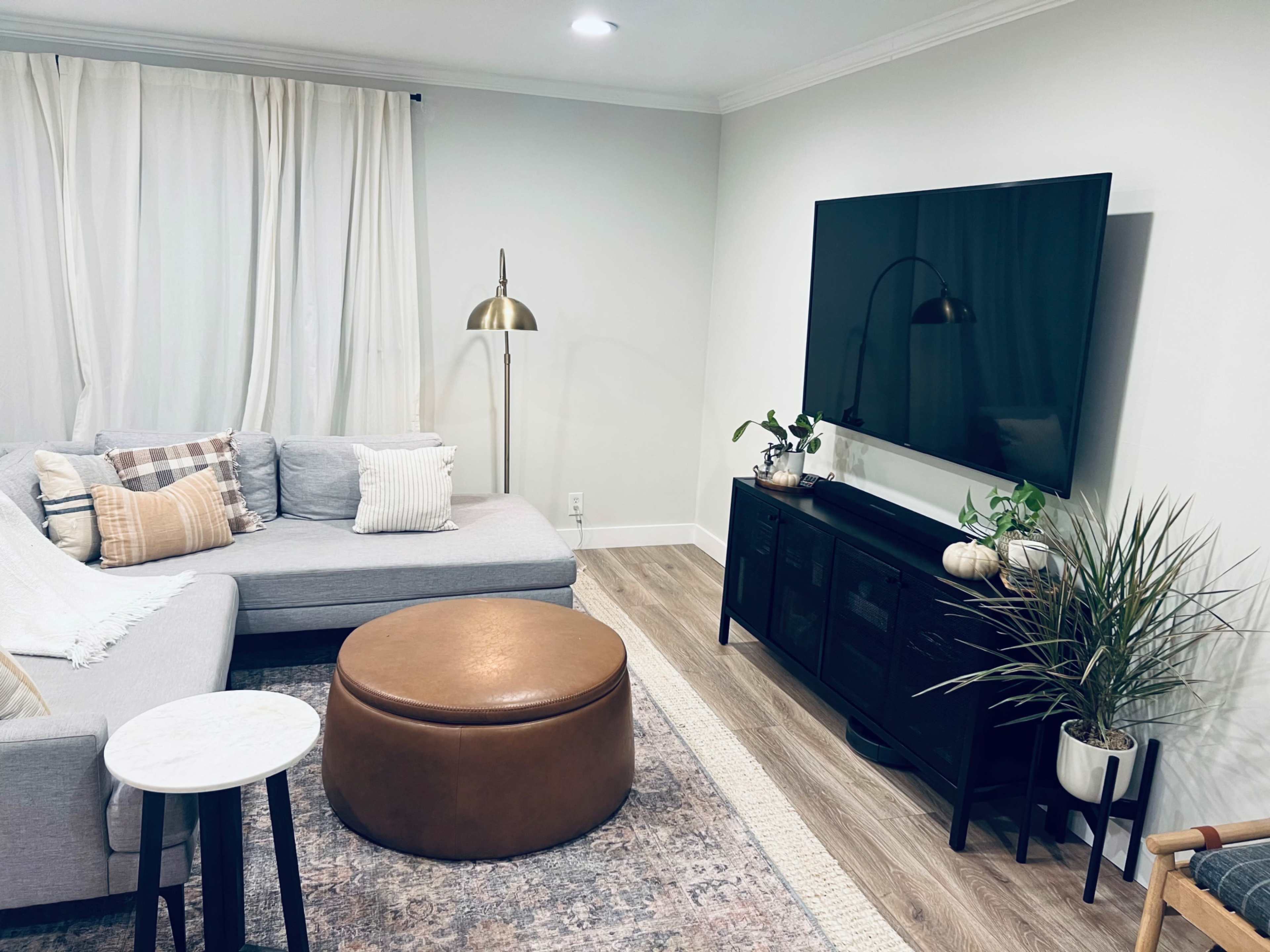 A modern living room features a gray sectional sofa, an ottoman, a wall-mounted television, and indoor plants arranged around the space.