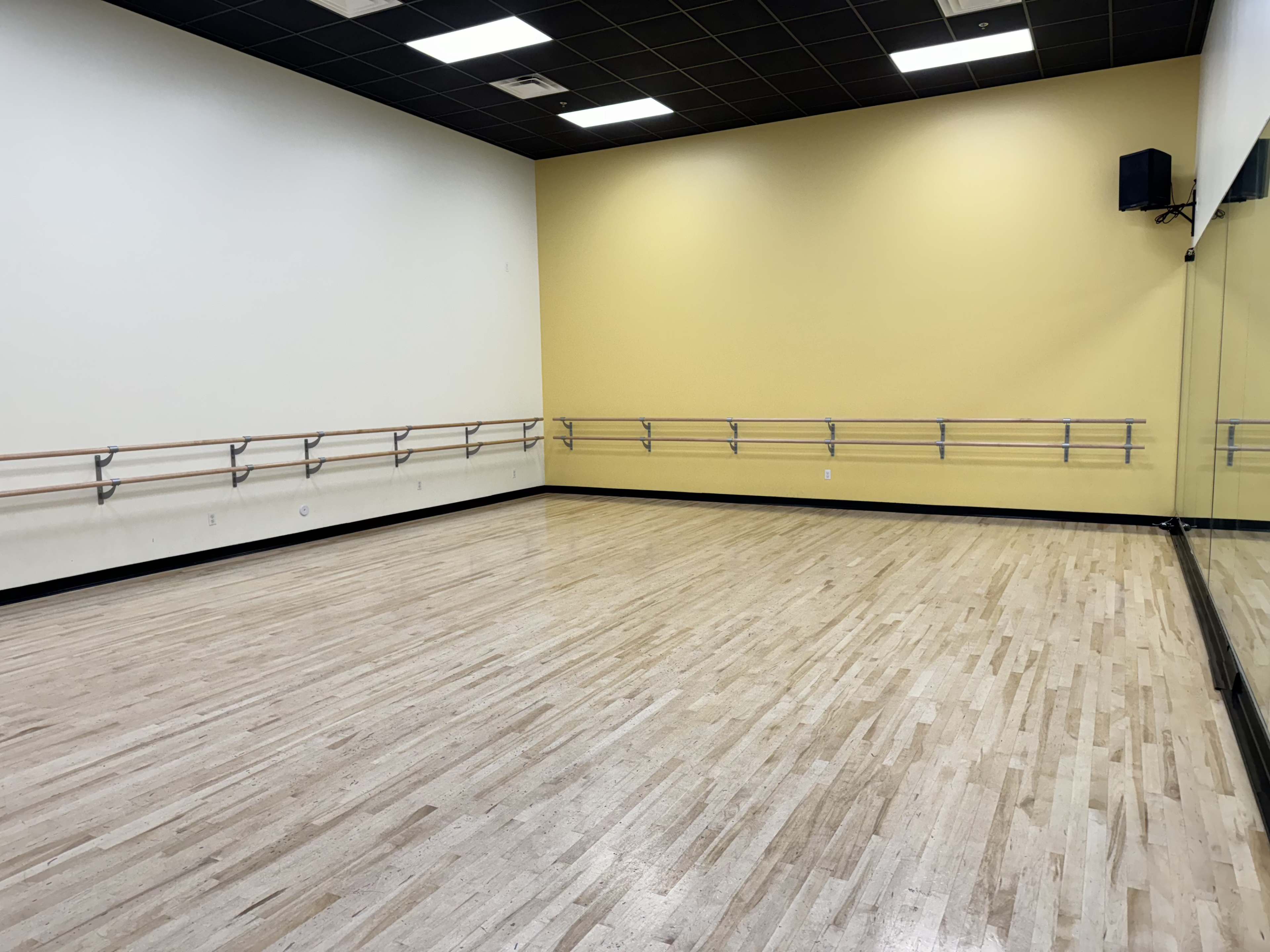 An empty dance studio with wooden flooring, yellow walls, and mirrored panels along one side.