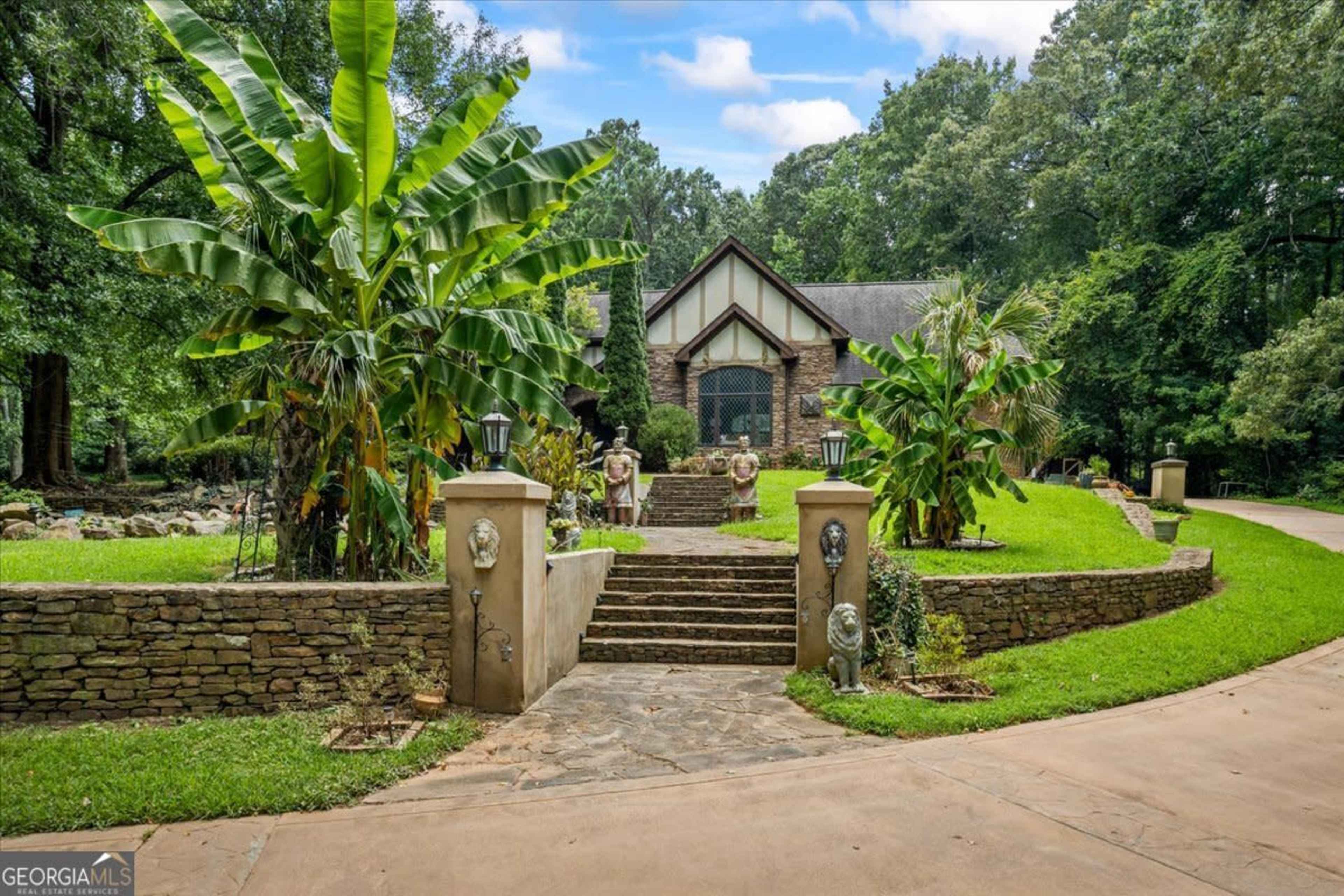 A stone-faced house sits at the end of a paved driveway, surrounded by lush greenery and decorative statues.