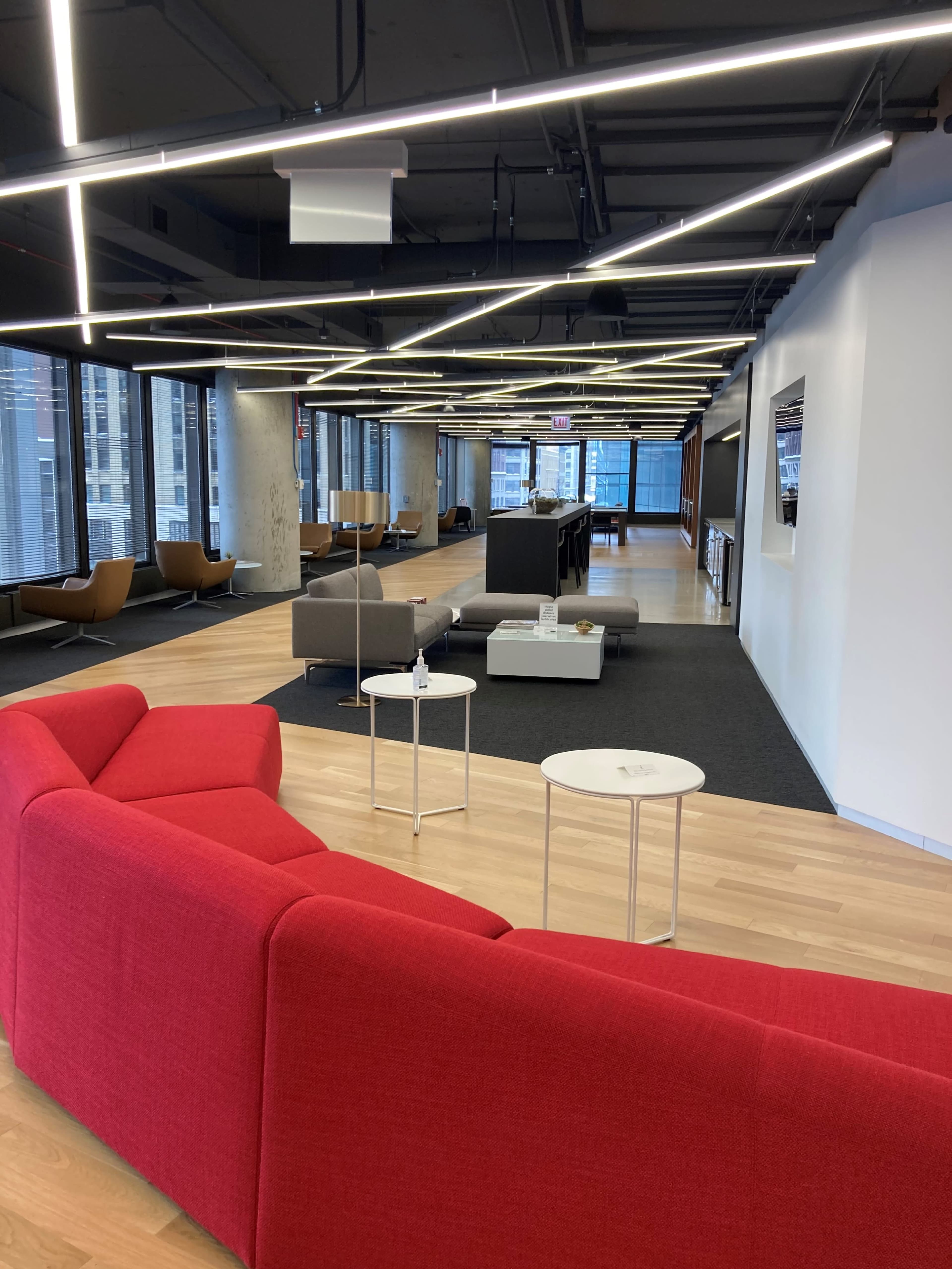 The image shows a spacious modern office interior featuring a red sectional sofa, various seating areas, and large windows with city views.