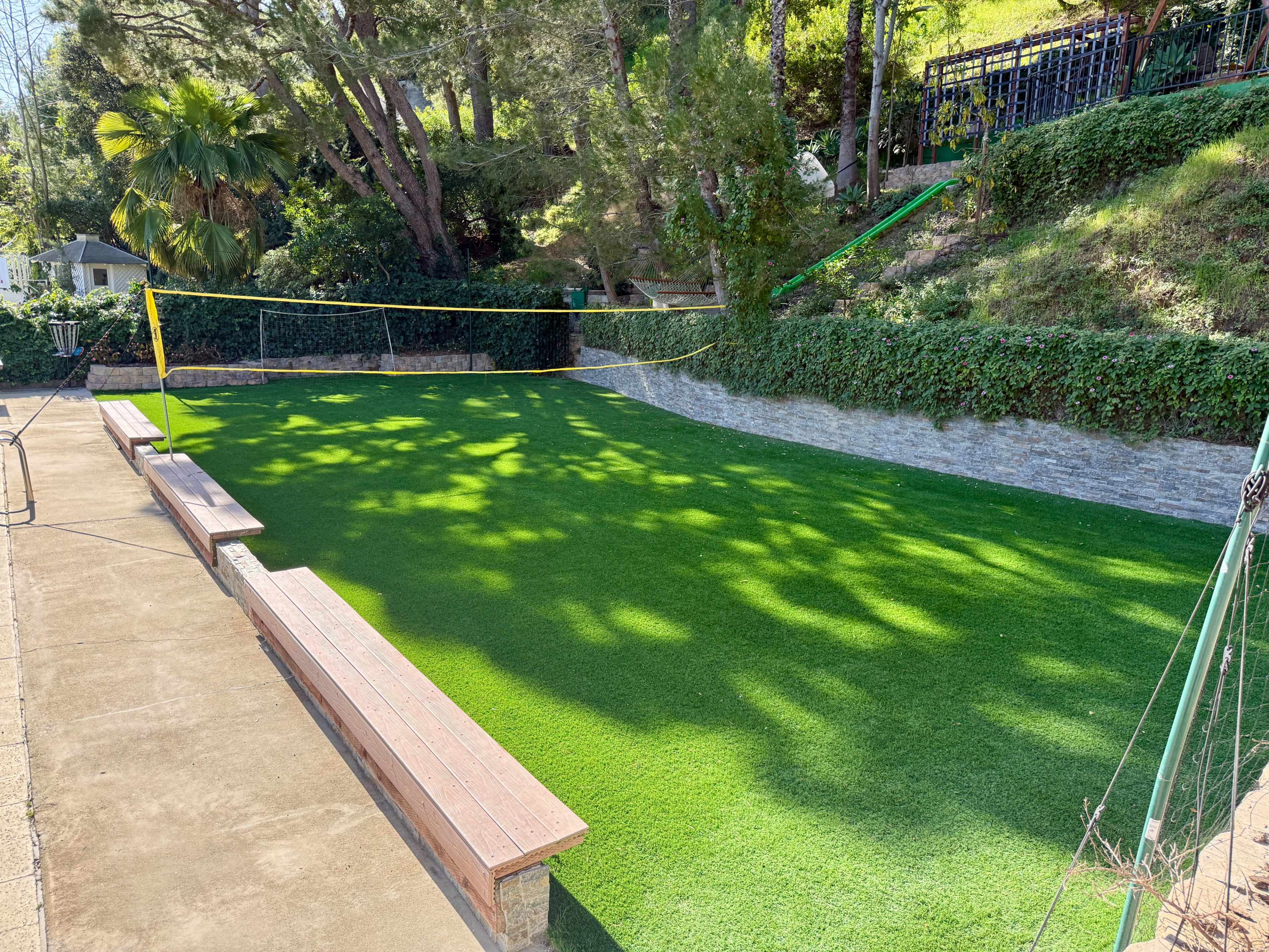 The image shows a green, artificial turf area designed for playing volleyball, bordered by stone walls and surrounded by trees.