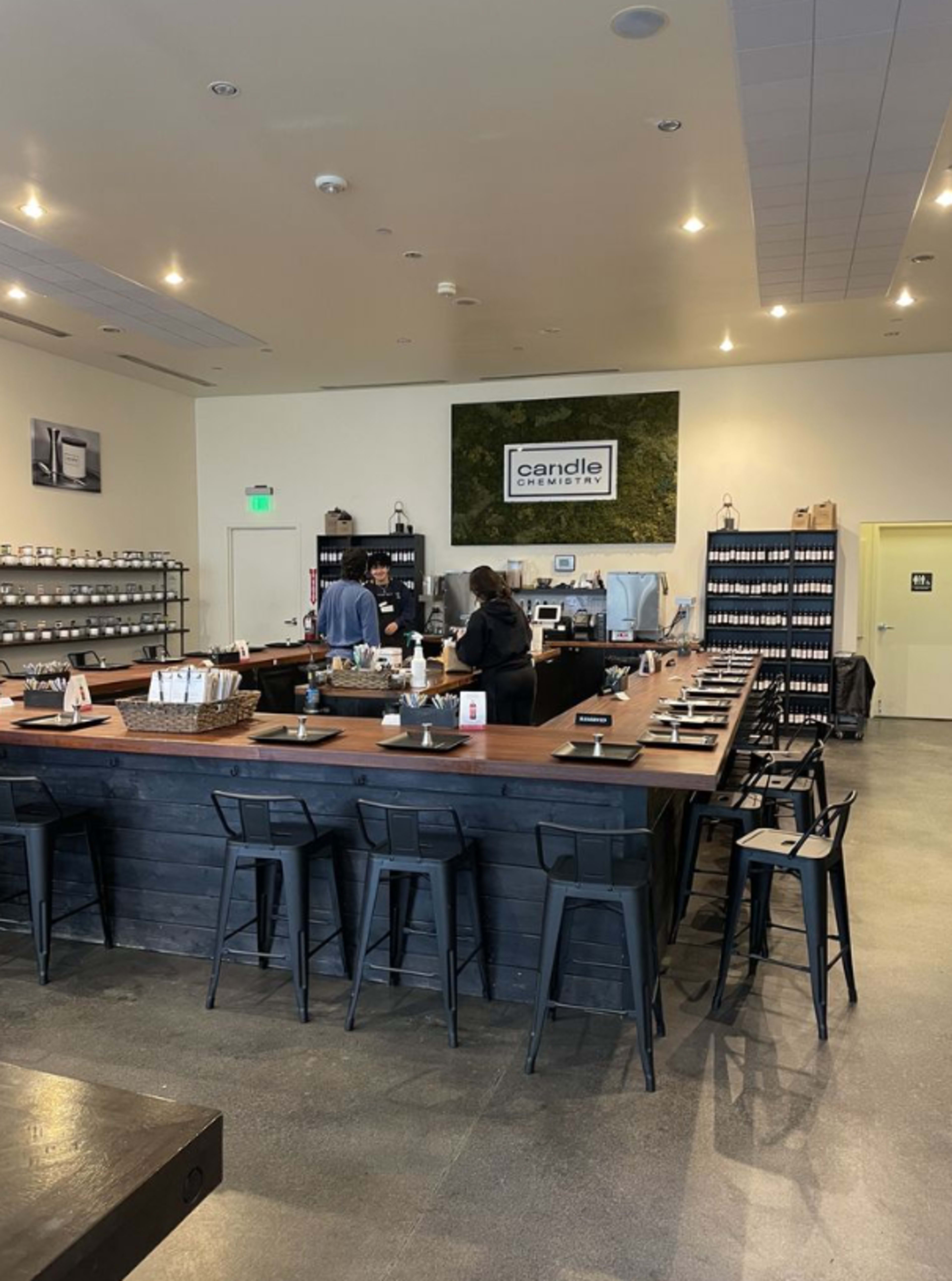 The image shows a modern candle-making shop with a long dark wood counter, bar stools, shelves lined with various candle jars, and two employees engaged in conversation.
