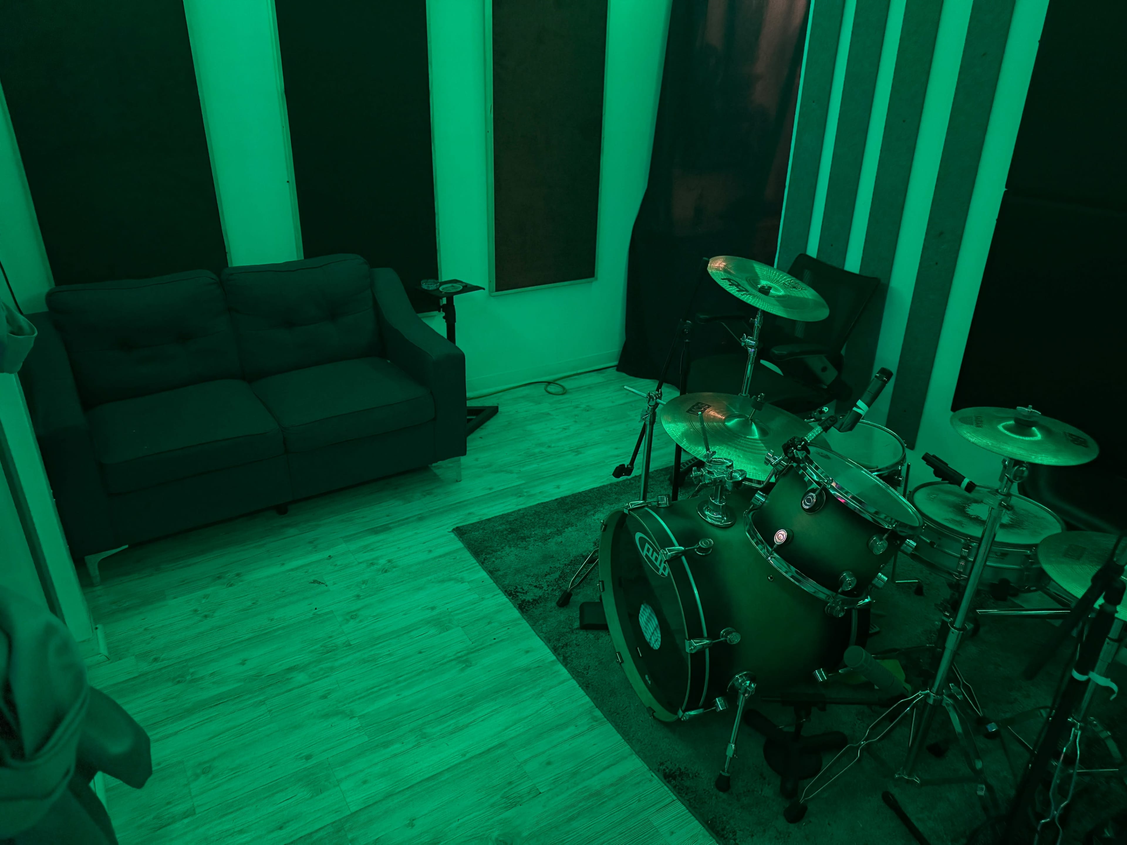 The image shows a small music studio featuring a drum kit on a rug beside a couch in a green-tinted room.
