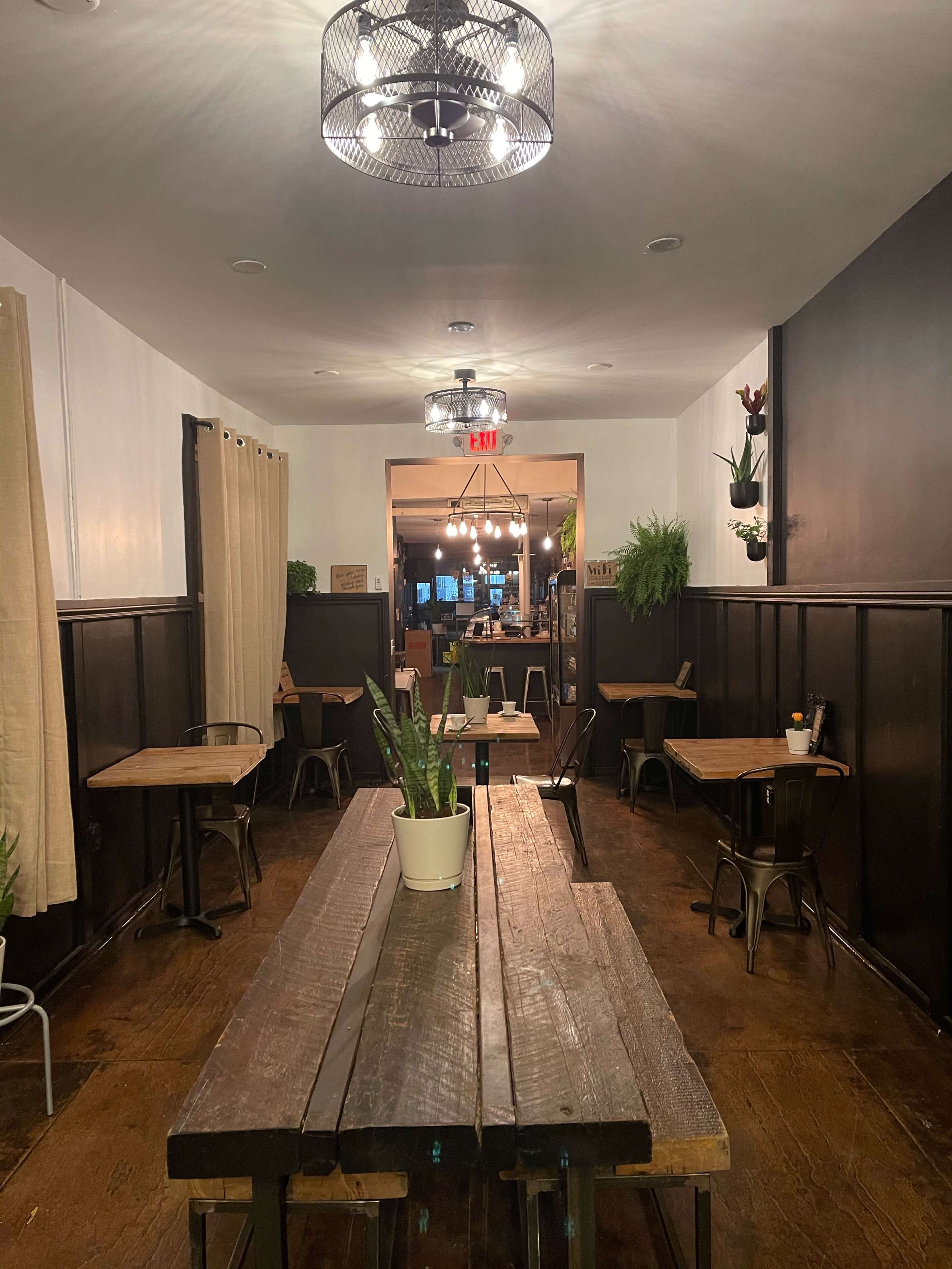 A narrow, dimly-lit restaurant hallway with wooden tables, plants, and a visible entrance to the kitchen area at the end.
