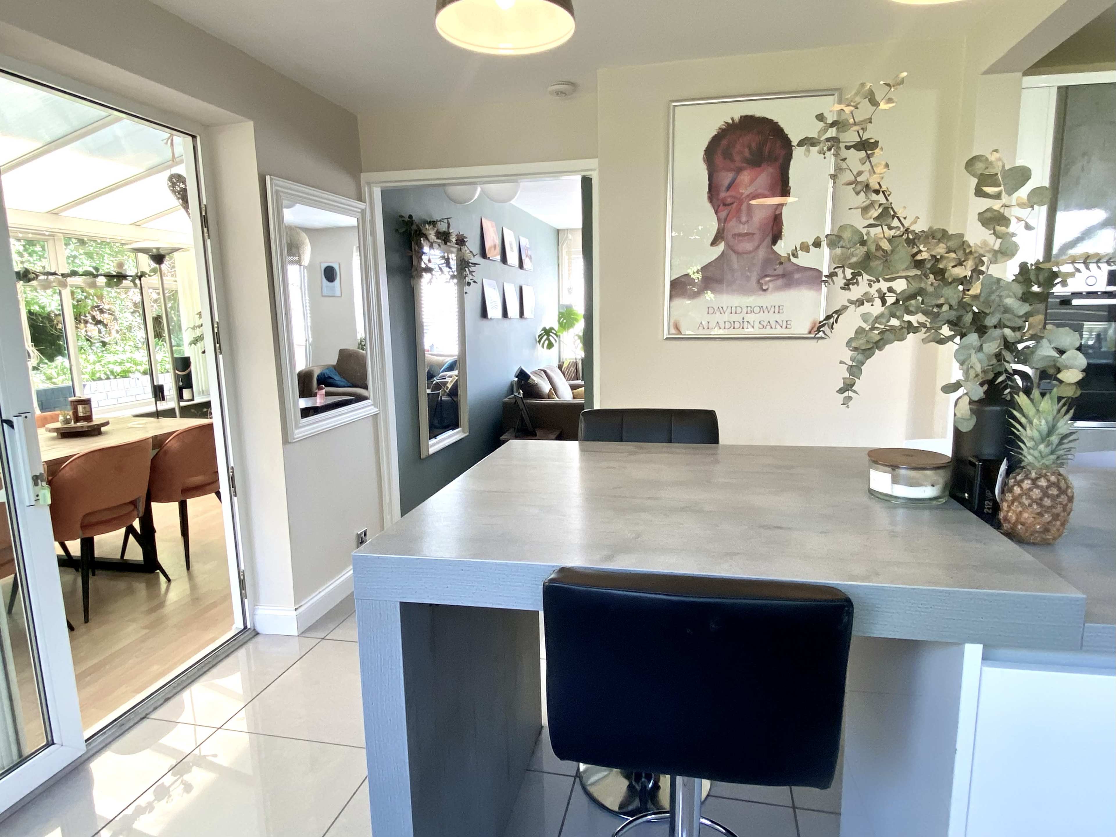 A modern kitchen area with a bar-style countertop, a poster of David Bowie on the wall, and a view of a sunlit room with seating in the background.