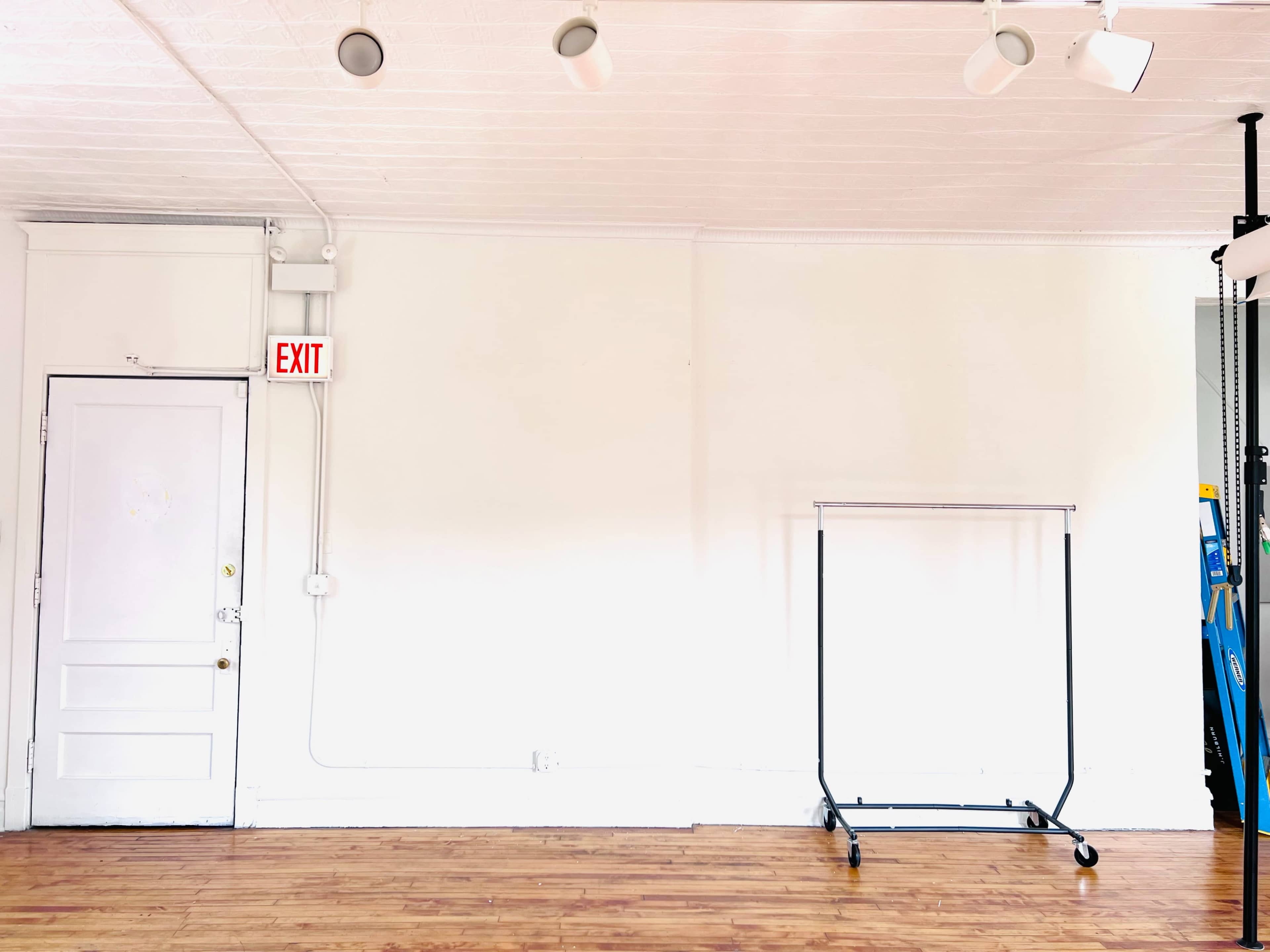 The image shows a bright, empty room with a white wall, a door marked with an exit sign, and a clothing rack positioned to the right.