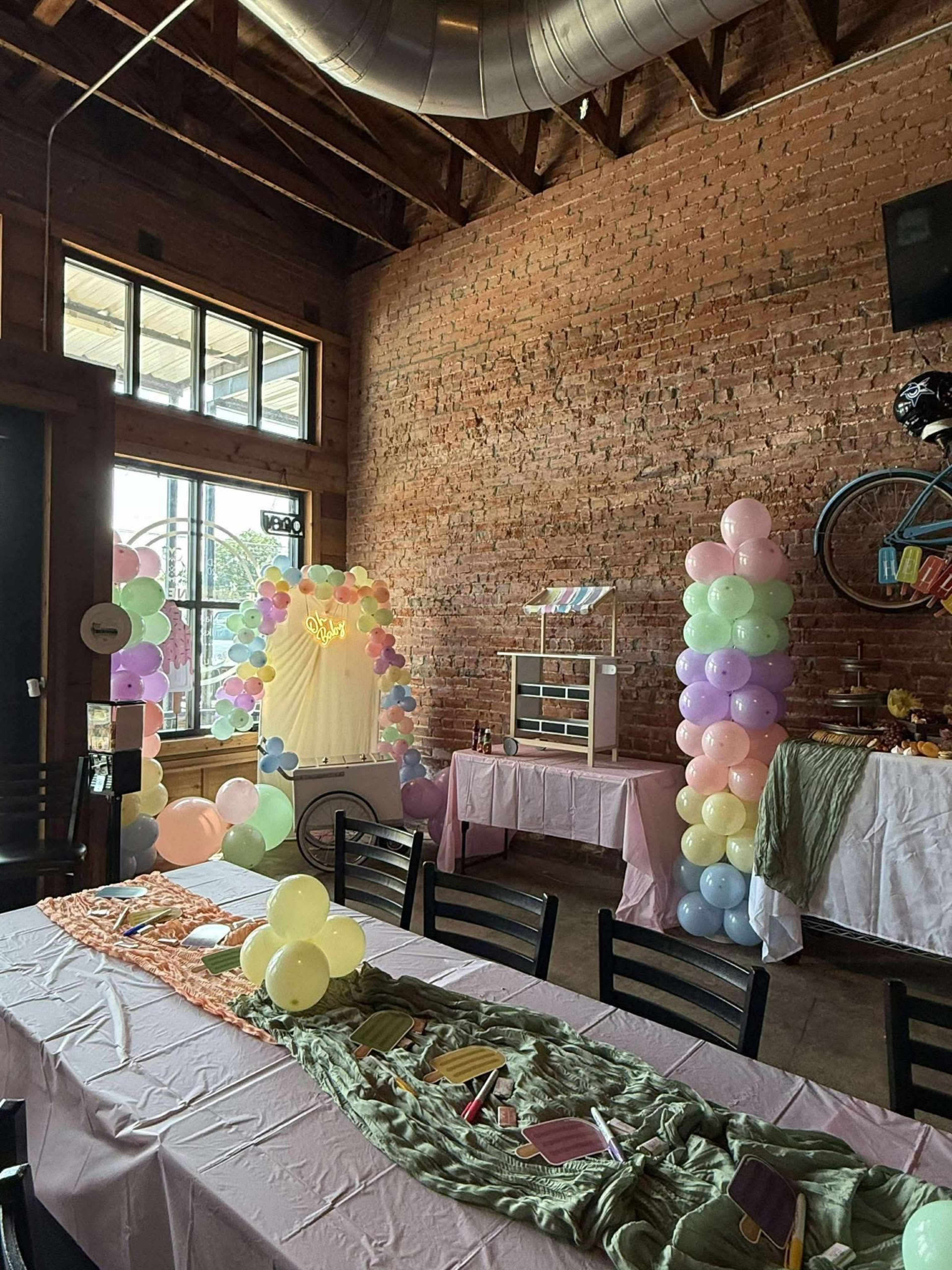 The image shows an event space adorned with pastel balloons, a decorative backdrop, and tables set for a gathering under exposed wooden beams.