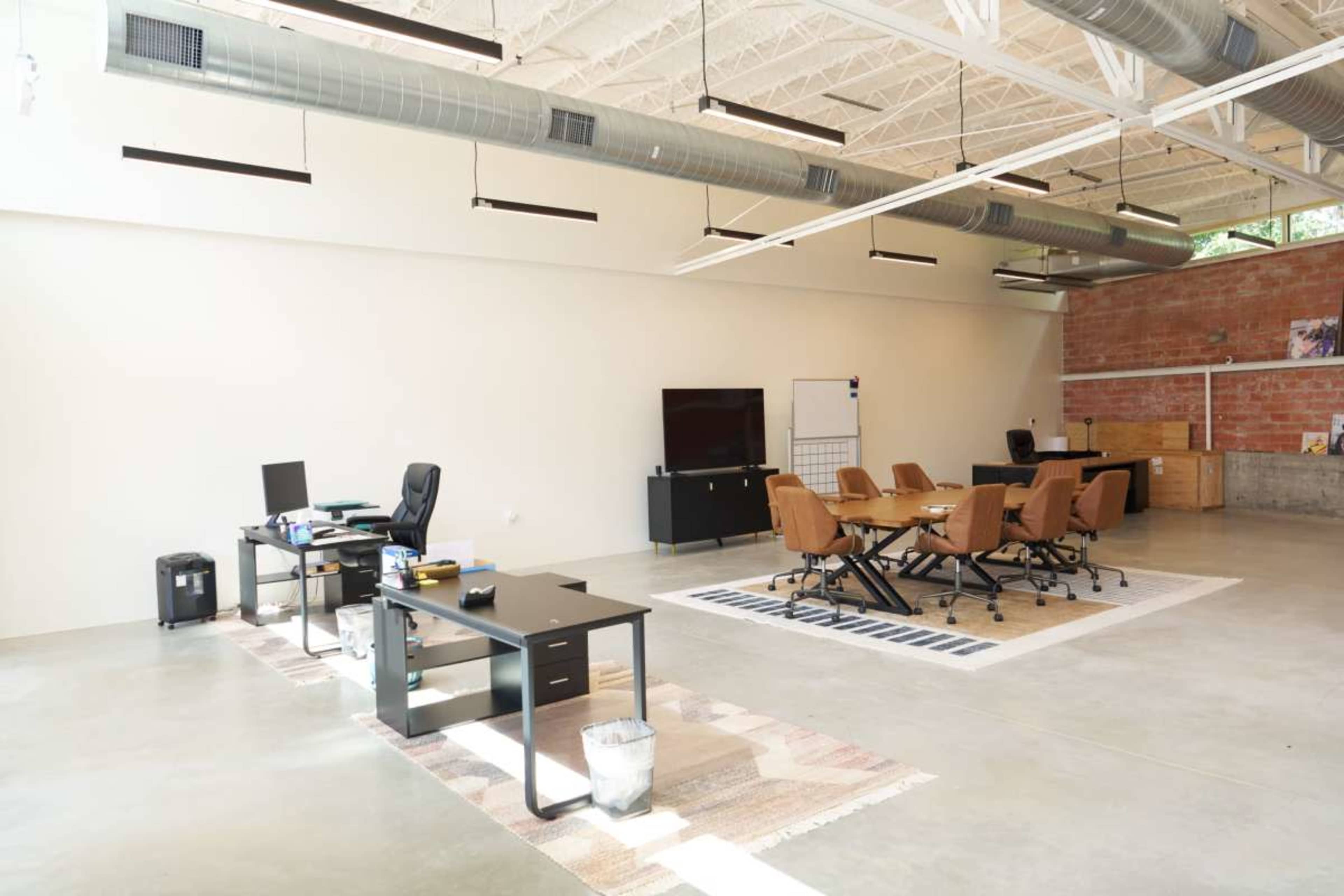 The image shows a modern office space with two desks, a conference table surrounded by chairs, and a large wall-mounted screen.