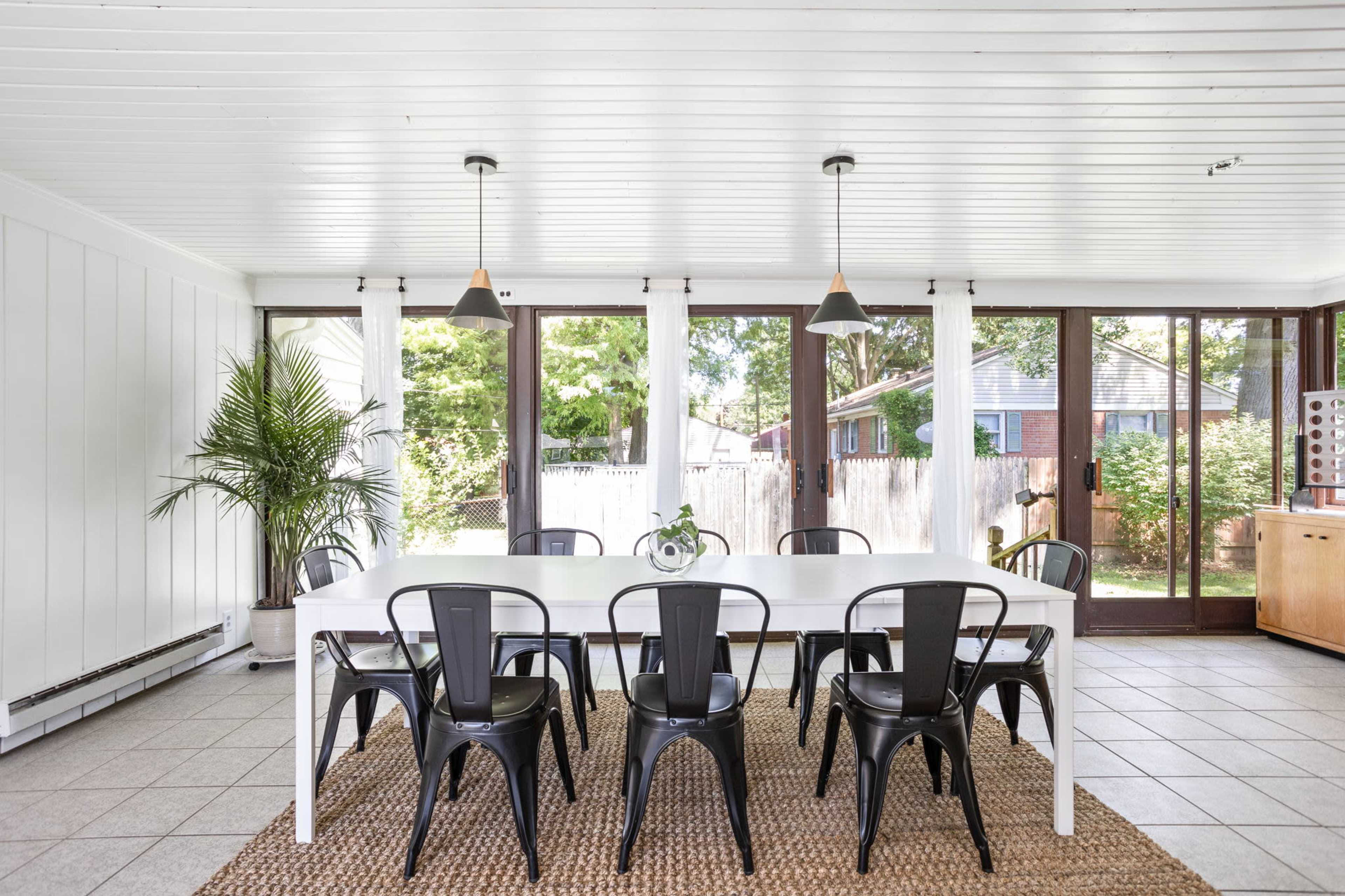 A light-filled dining area with a long white table surrounded by black chairs, large windows, and a view of a green backyard.