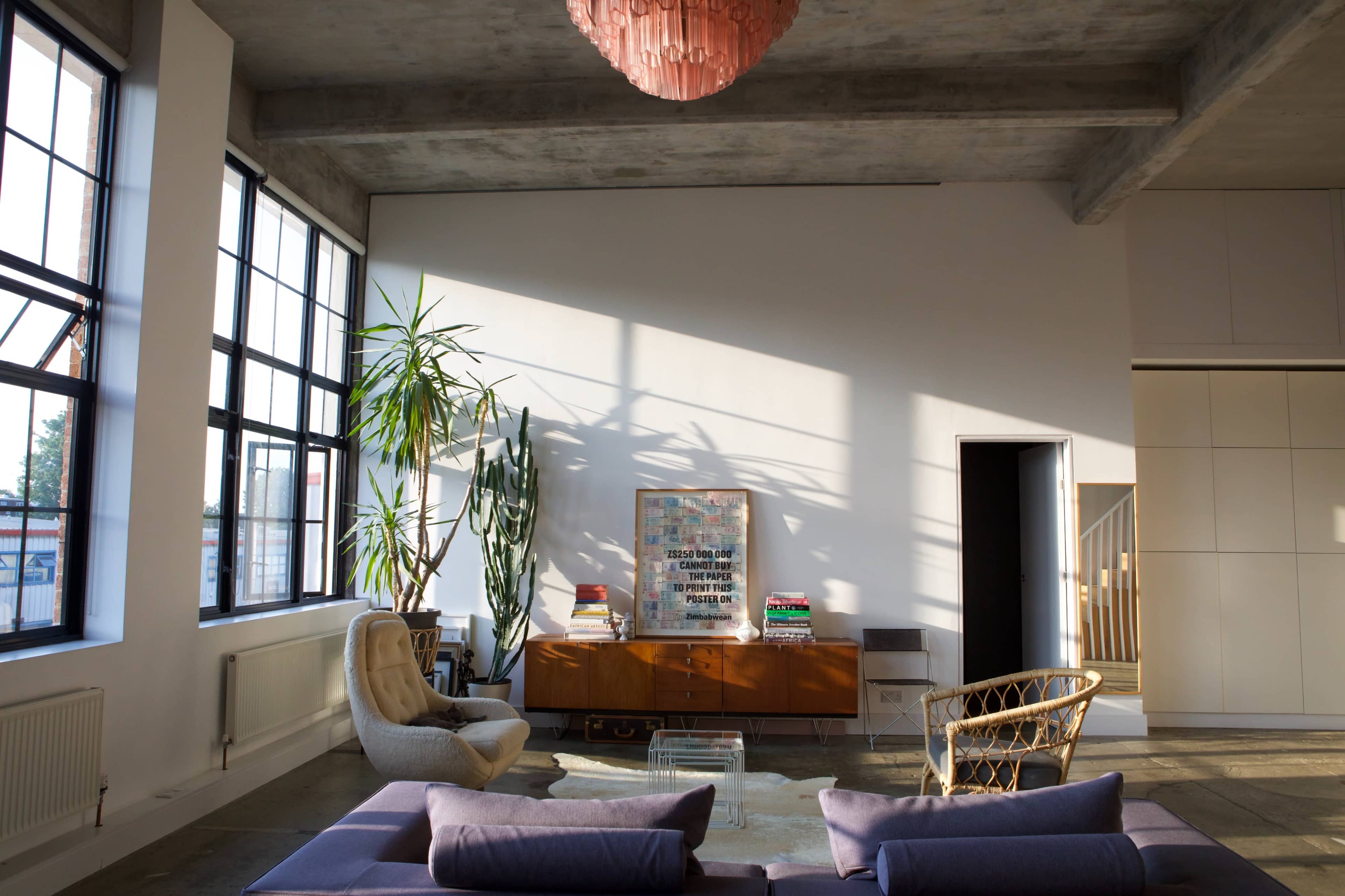The image shows a bright, modern living room with large windows, a mixture of plants, and minimalist furniture including a low sofa and a wooden cabinet.