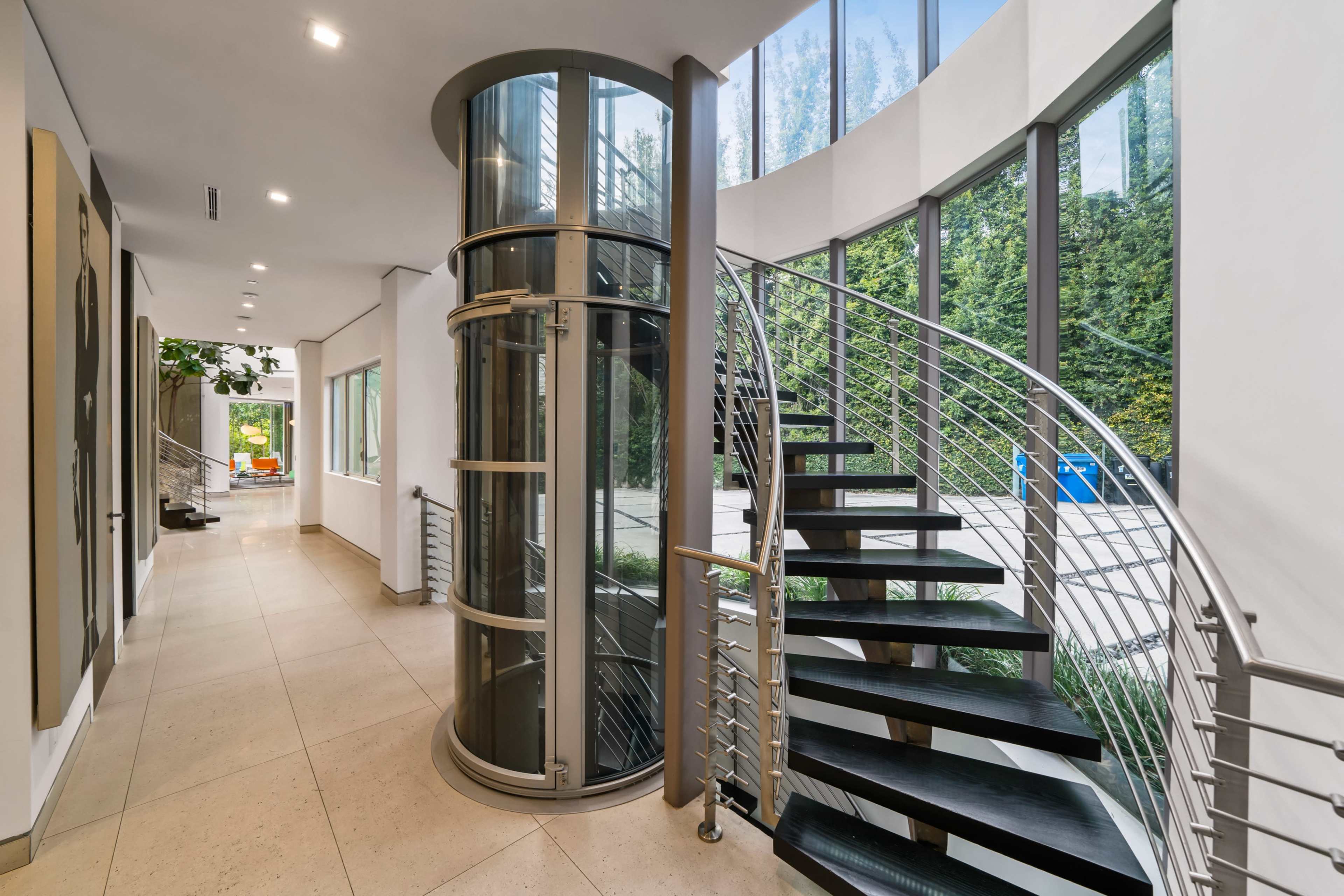 A modern interior with a curved staircase, a glass elevator shaft, and large windows overlooking greenery.
