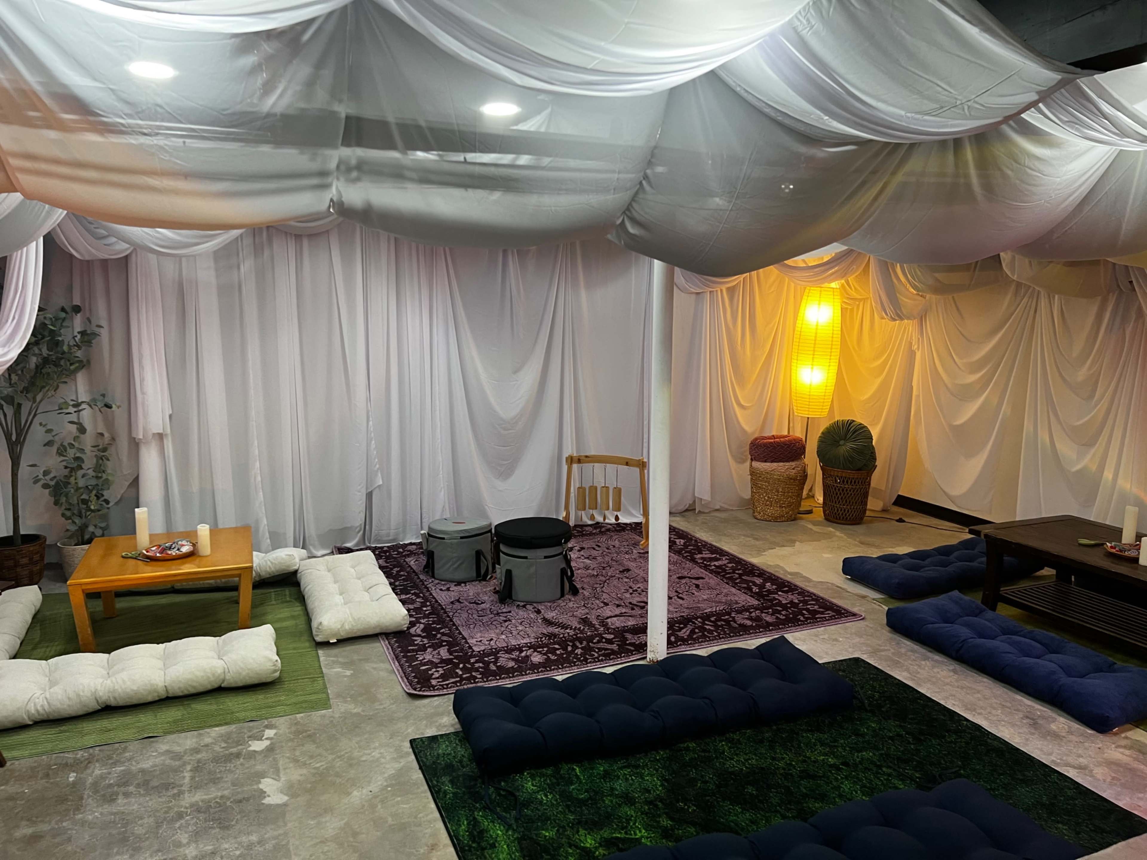 The image shows a spacious interior room with various seating arrangements, including cushions on the floor and tables, surrounded by draped fabric and soft lighting.