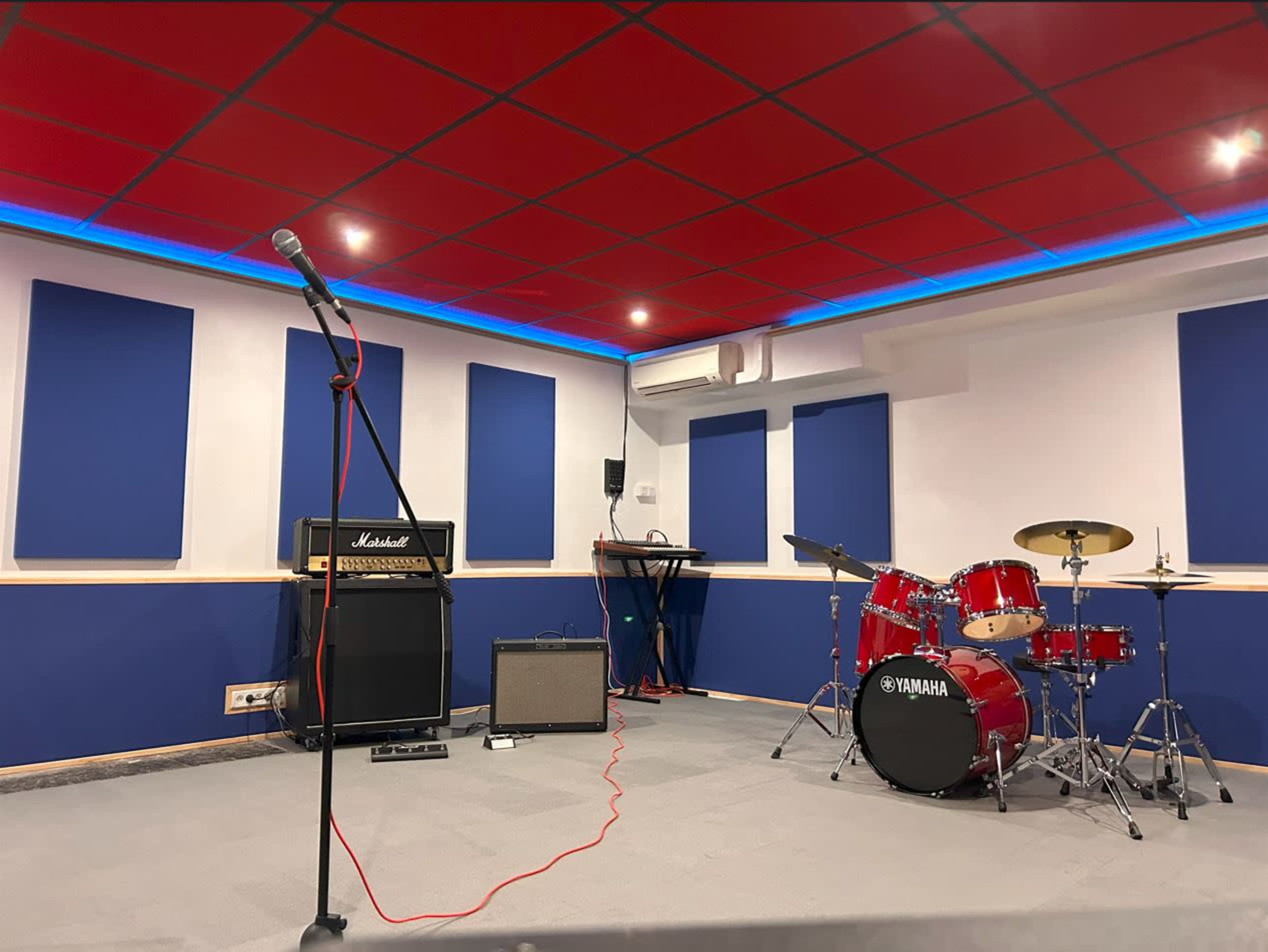 A music rehearsal studio with a microphone on a stand, an amplifier, a keyboard, and a red drum set.