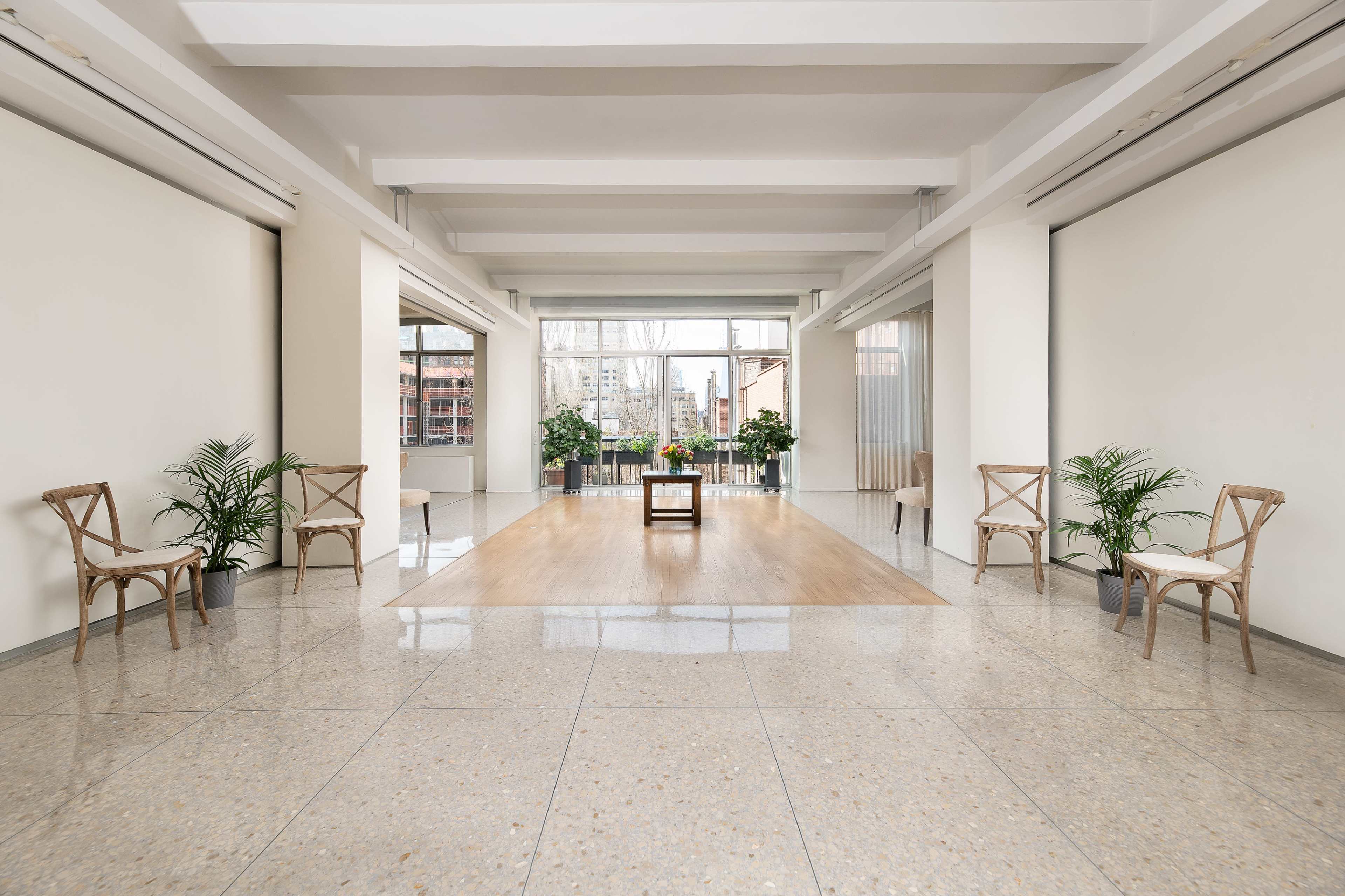 A spacious, minimalist room with wooden flooring, two plants in pots, a central table, and four wooden chairs arranged near the walls.