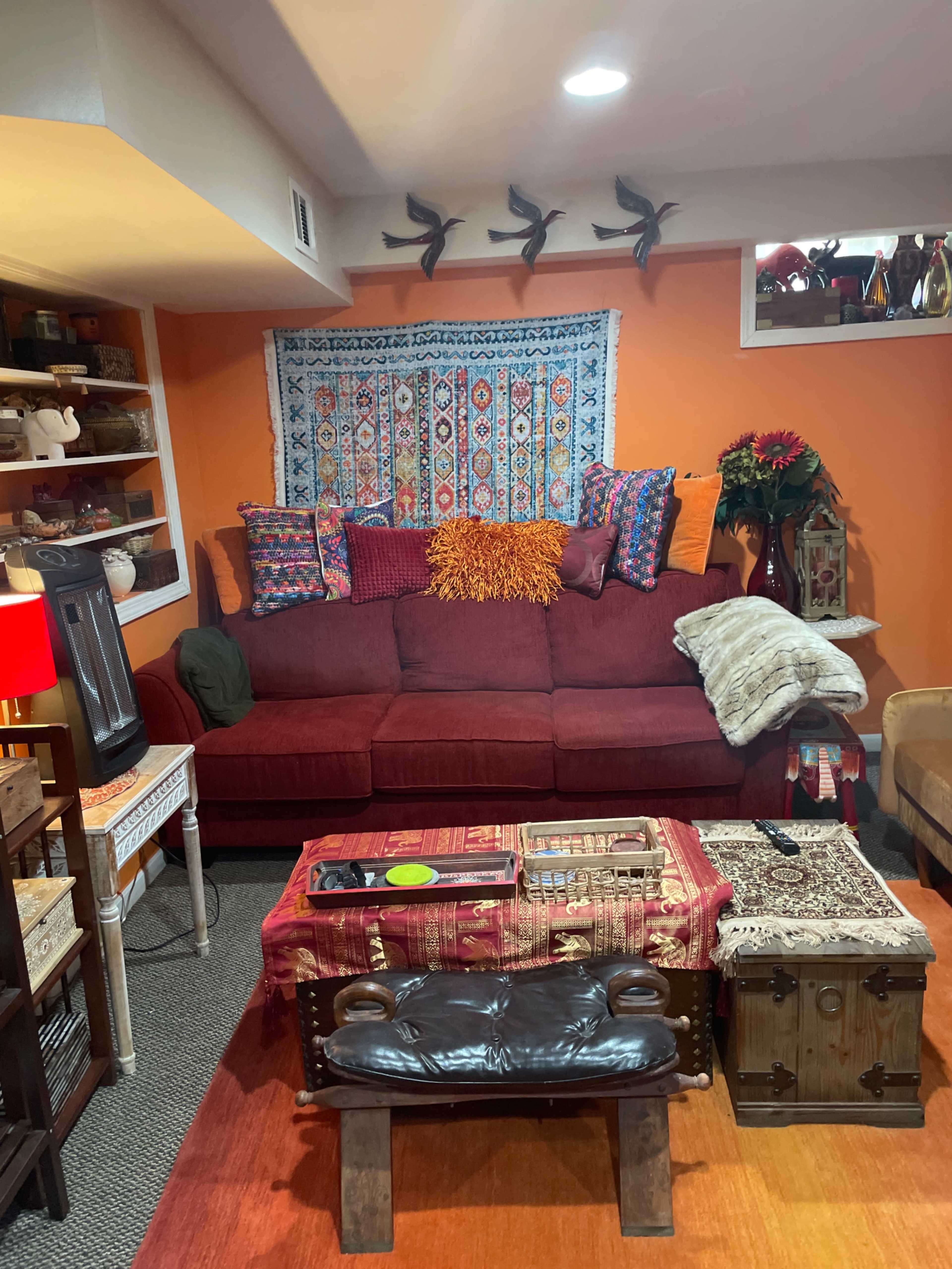 The image shows a cozy living room with a red couch adorned with colorful cushions, a patterned wall tapestry, and various decorative items on shelves.