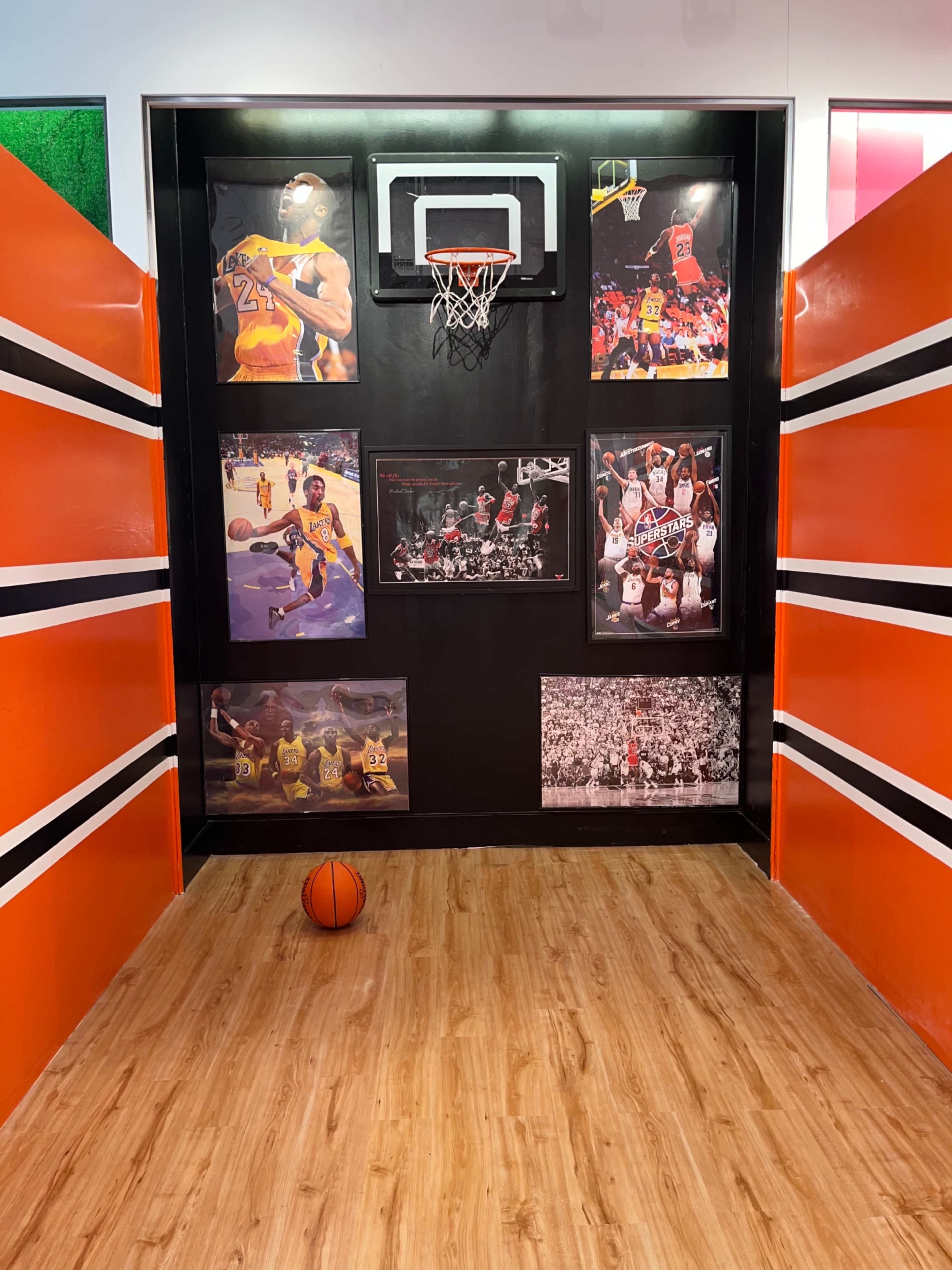 A small basketball court setup with a hoop on the wall, surrounded by framed photographs of basketball players, and a basketball on the wooden floor.