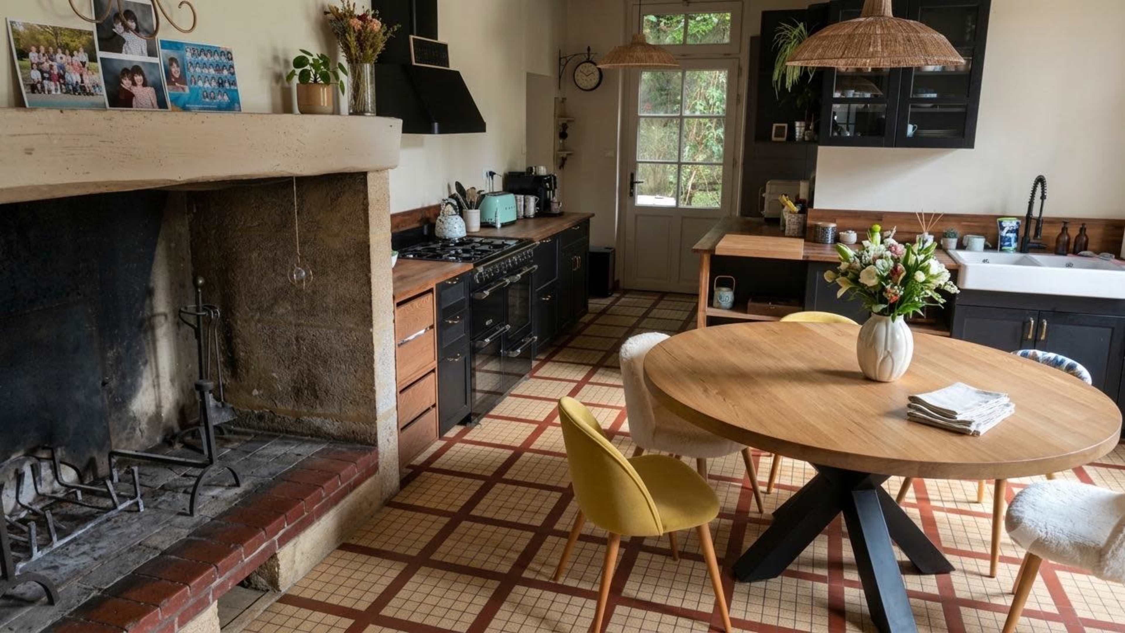 The image shows a modern kitchen with a circular wooden dining table, a stone fireplace, and black cabinetry, featuring a large window that opens to an outdoor view.