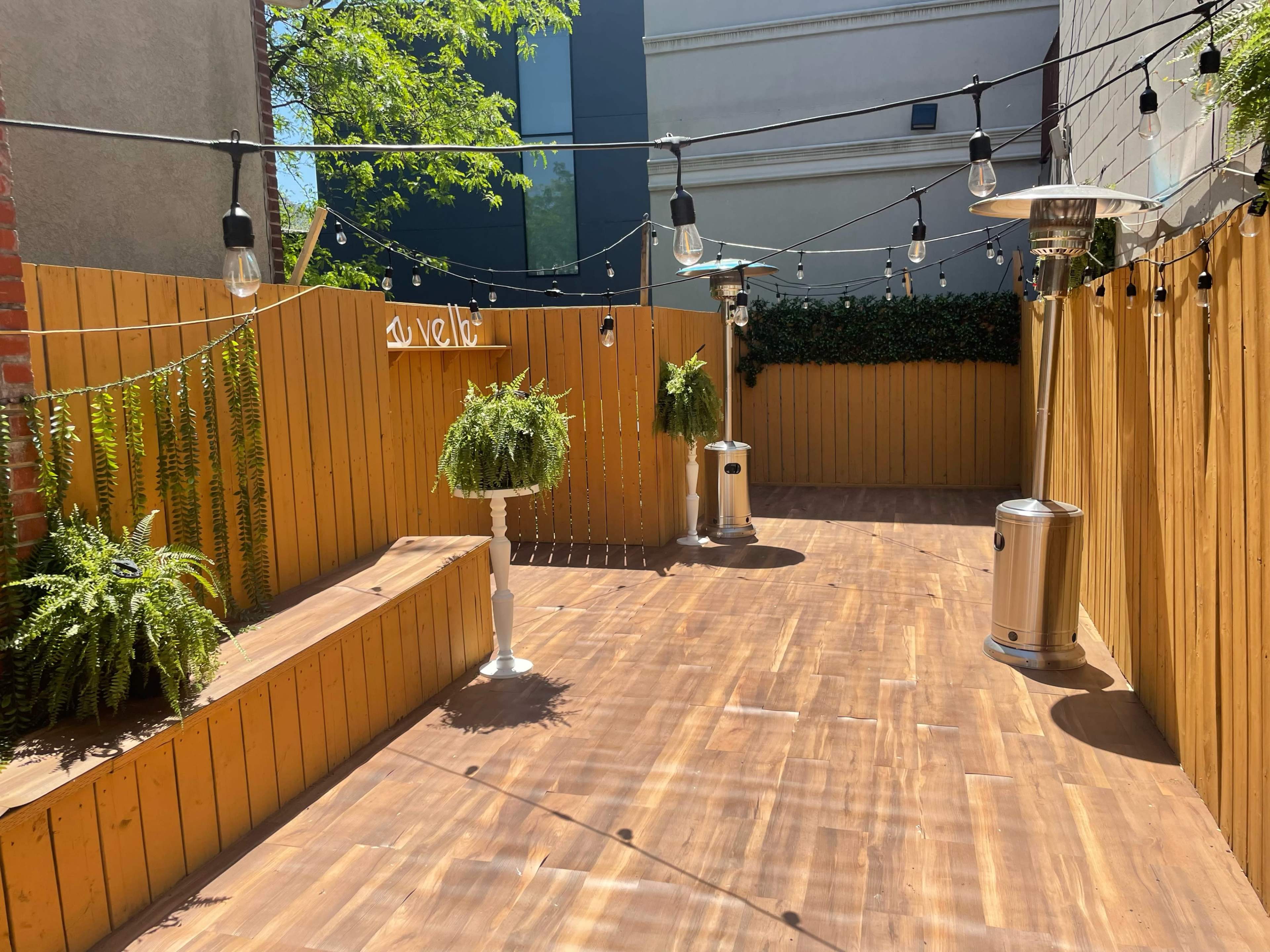 A wooden outdoor space with hanging lights, potted ferns, and patio heaters surrounded by wooden fencing.