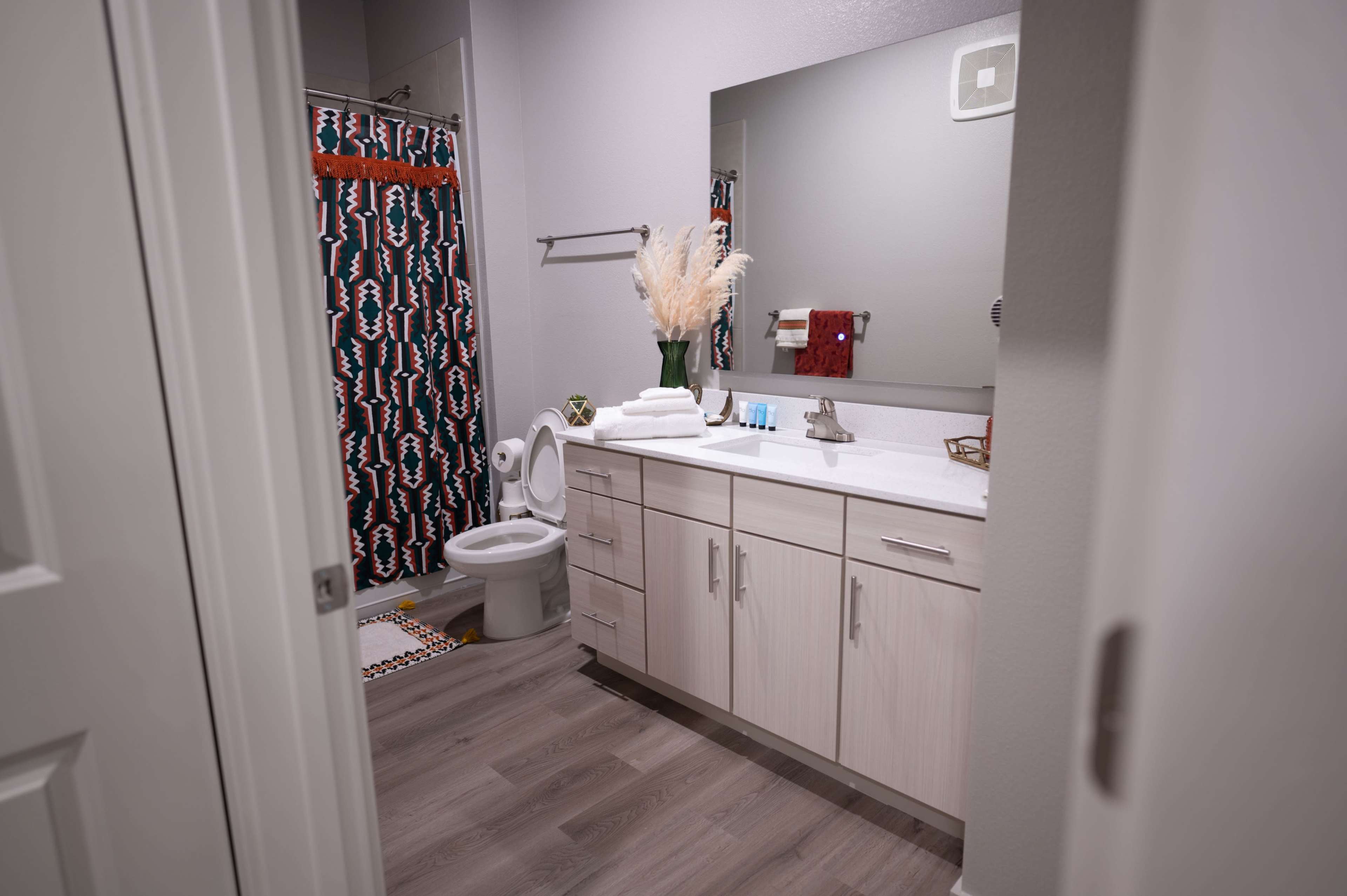 A modern bathroom features a white countertop, a mirror, a toilet, and a shower curtain with a colorful geometric pattern.