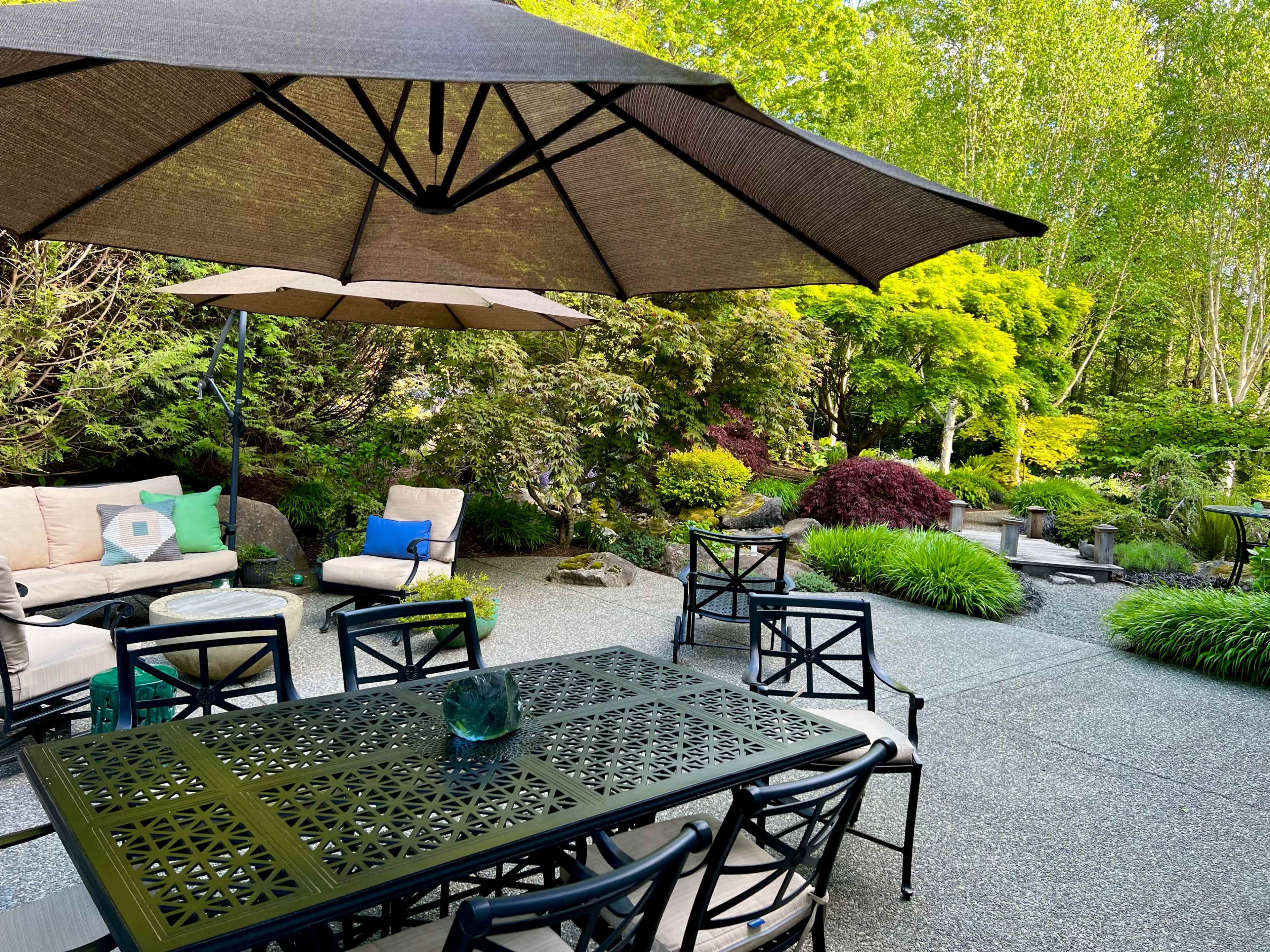 A backyard patio with a table and chairs under a large umbrella, surrounded by lush greenery and decorative plants.