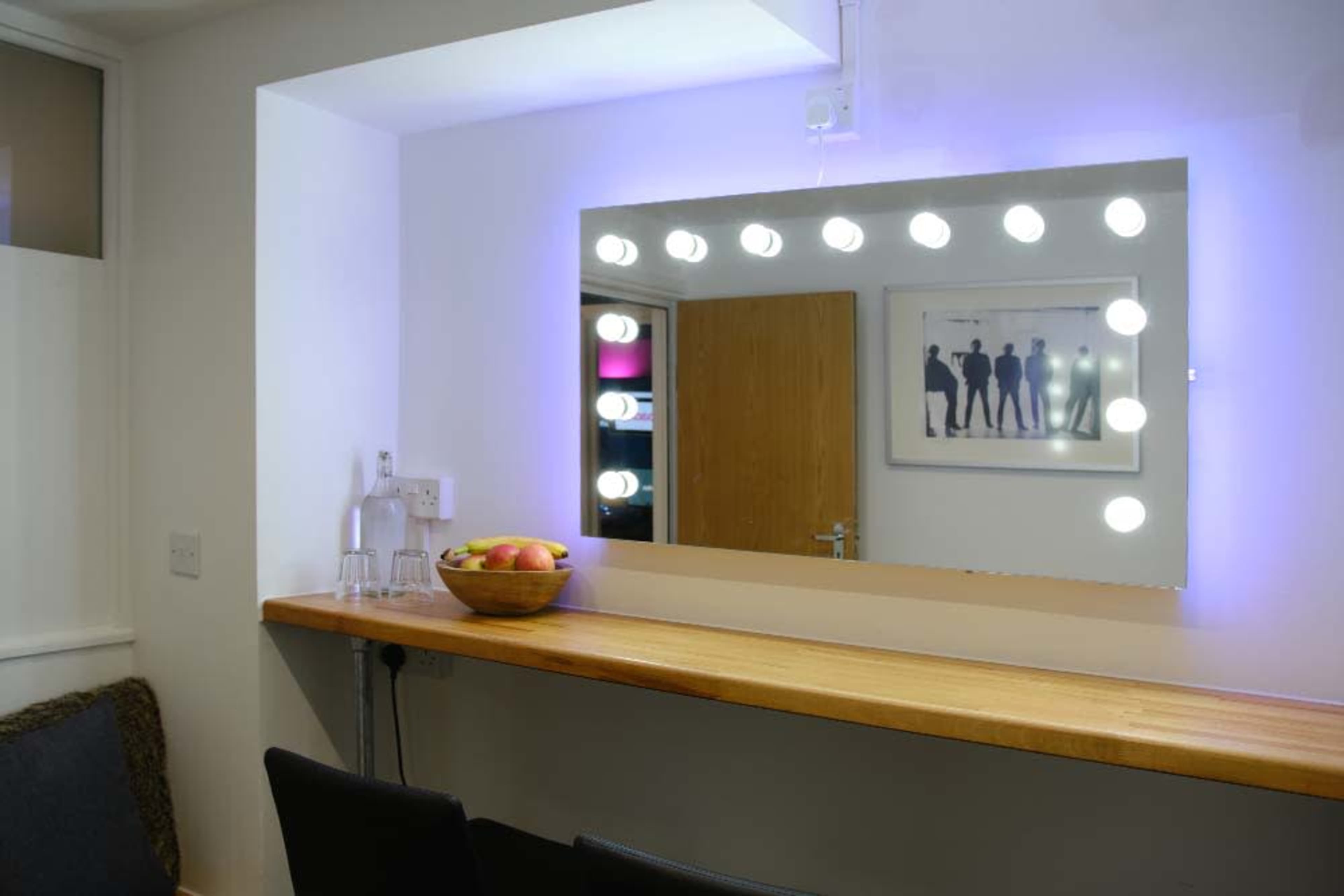 A mirror with circular lights is mounted on a wall above a wooden counter with a bowl of apples and a water pitcher nearby.