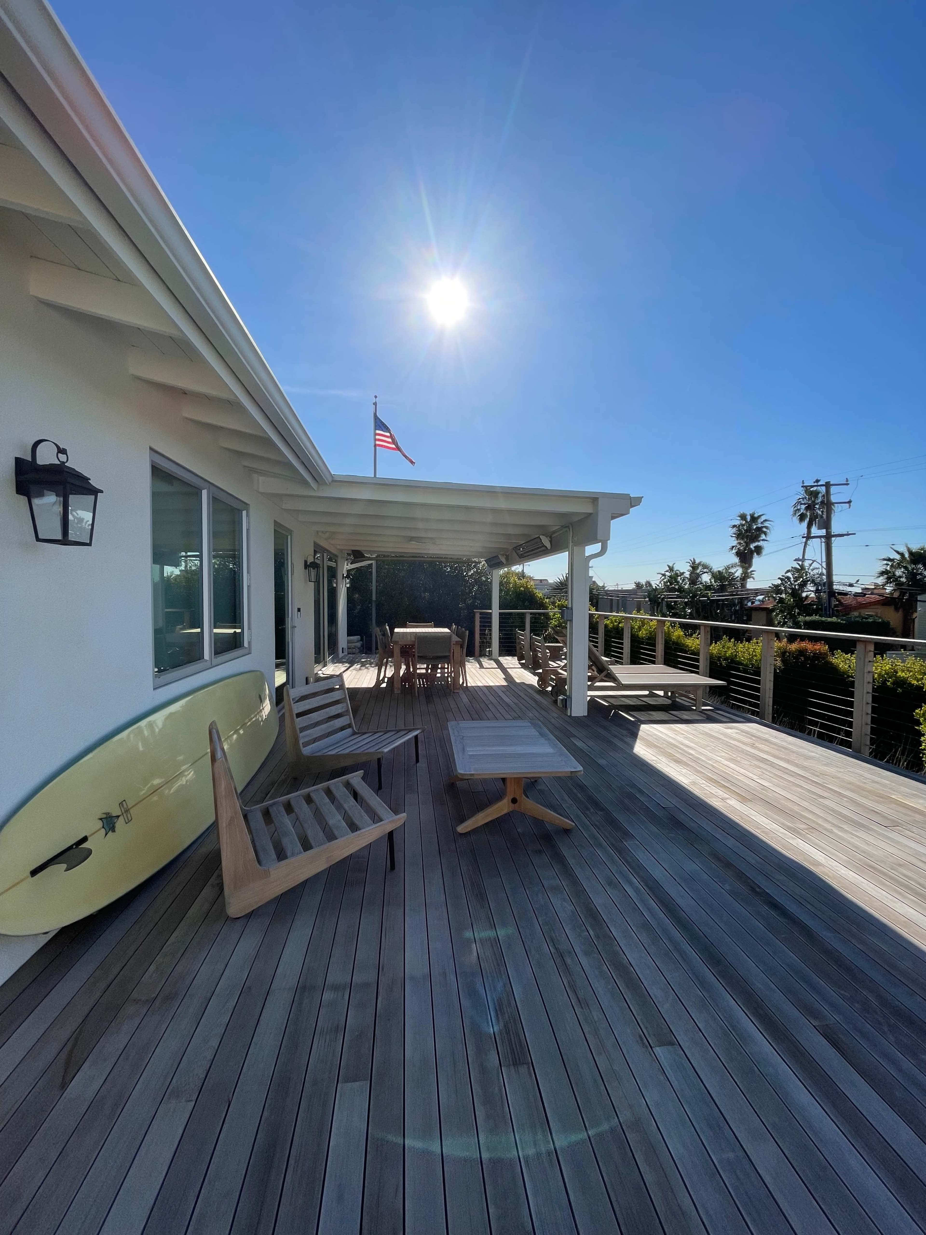A sunlit wooden deck with a surfboard leaning against the house, outdoor furniture, and an American flag waving in the background.