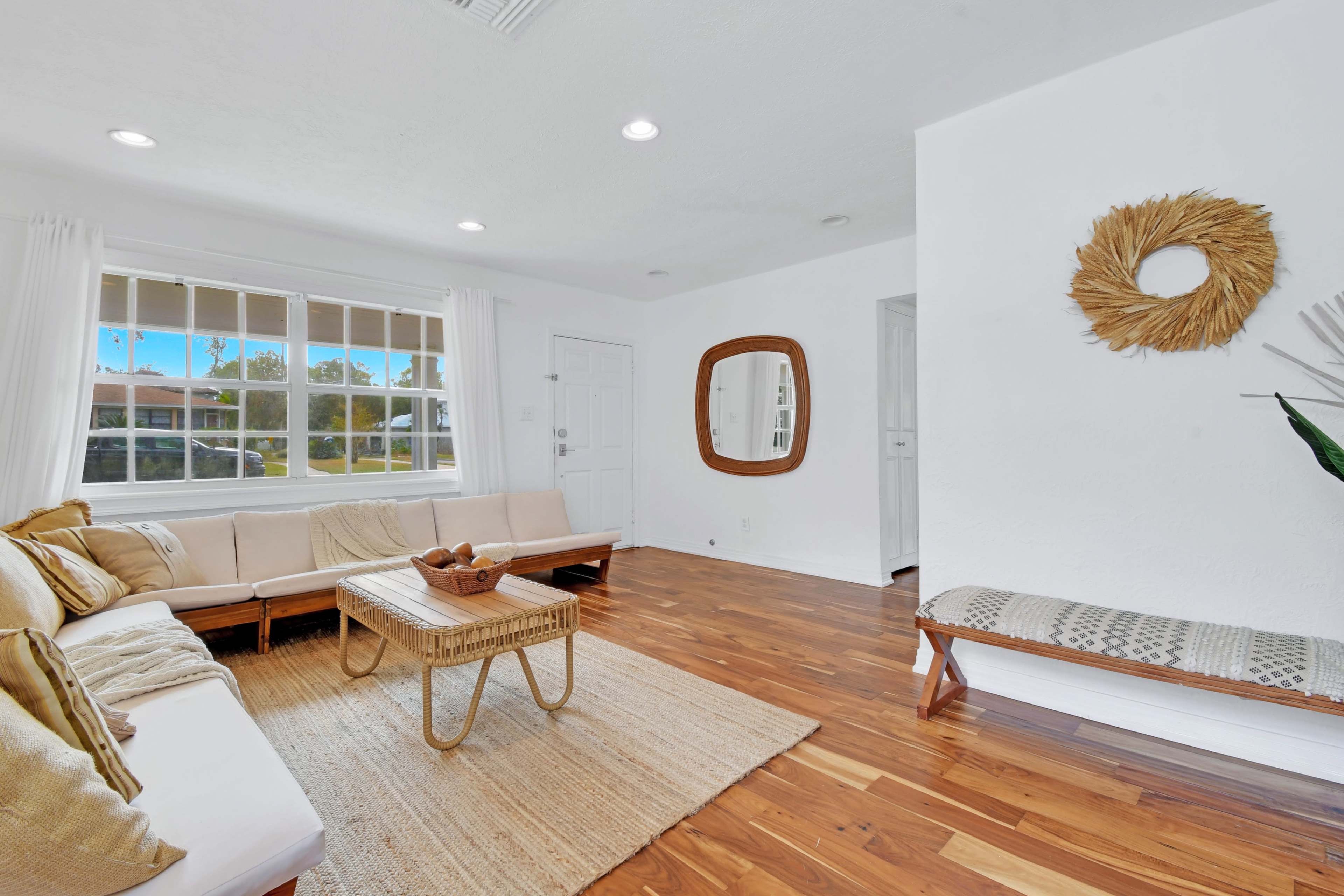 The image shows a bright living room with a large window, wooden flooring, a light-colored sofa, and a decorative mirror on the wall.
