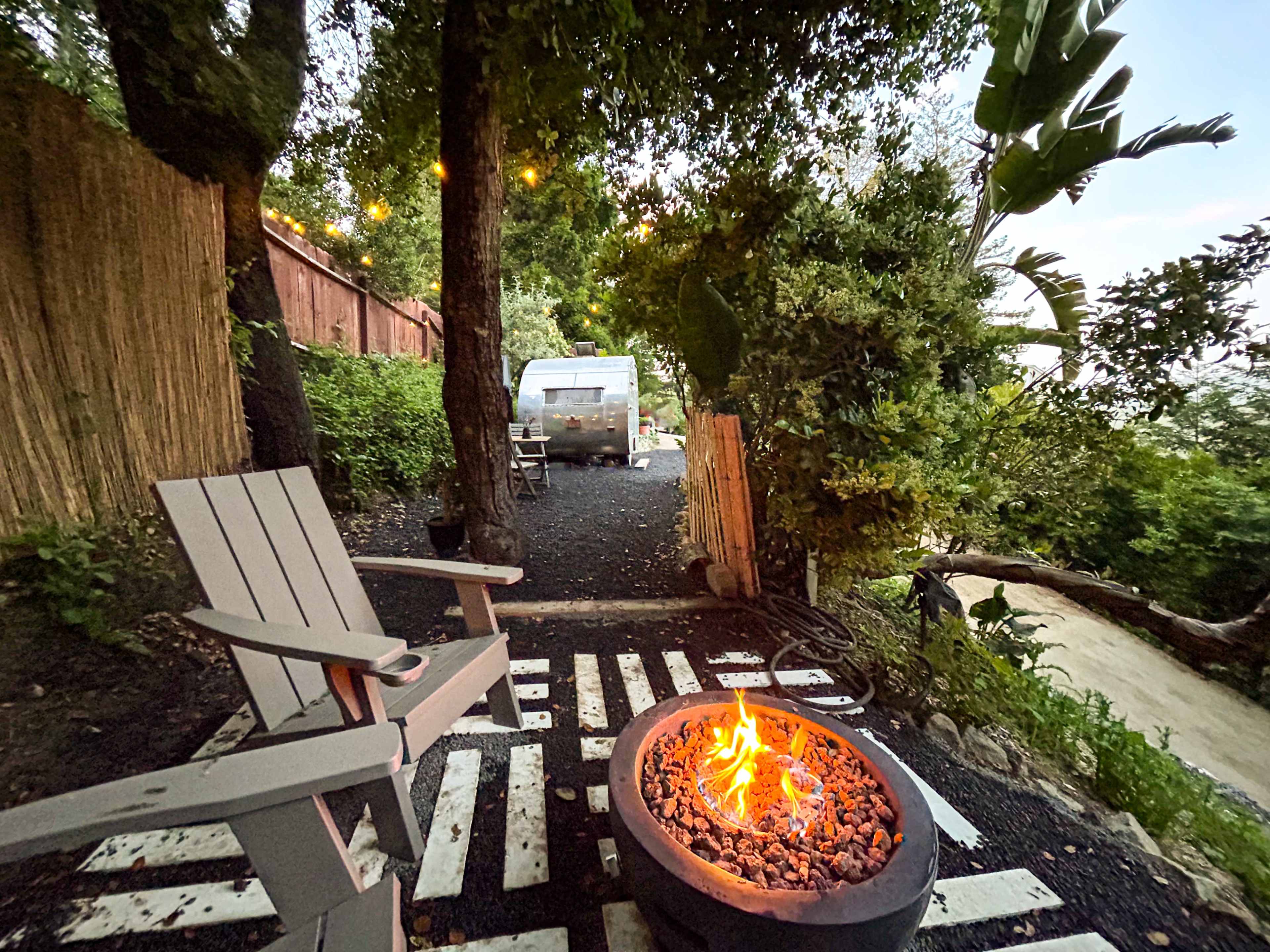 A cozy outdoor space features a fire pit surrounded by wooden chairs, with string lights illuminating a pathway leading to a parked Airstream trailer among lush greenery.
