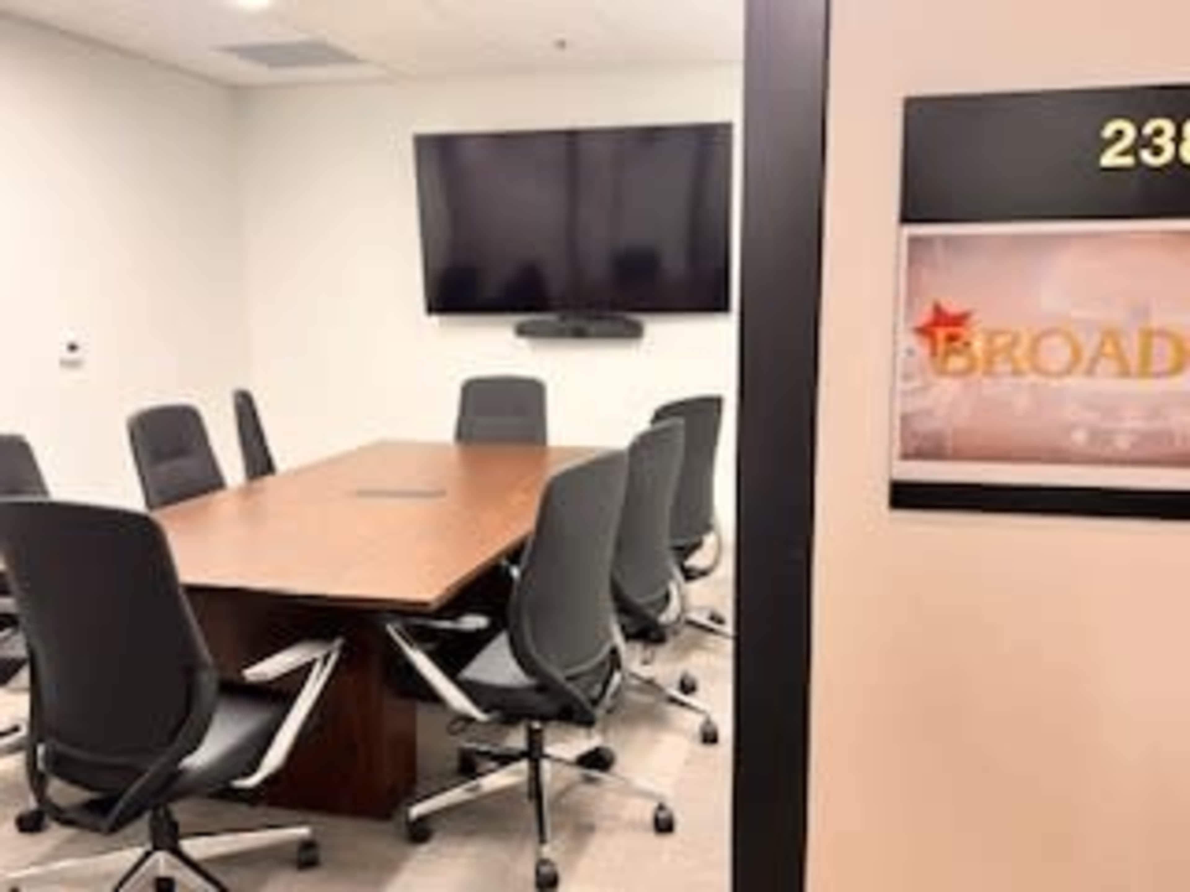 The image shows a conference room with a large table surrounded by six office chairs and a wall-mounted television.