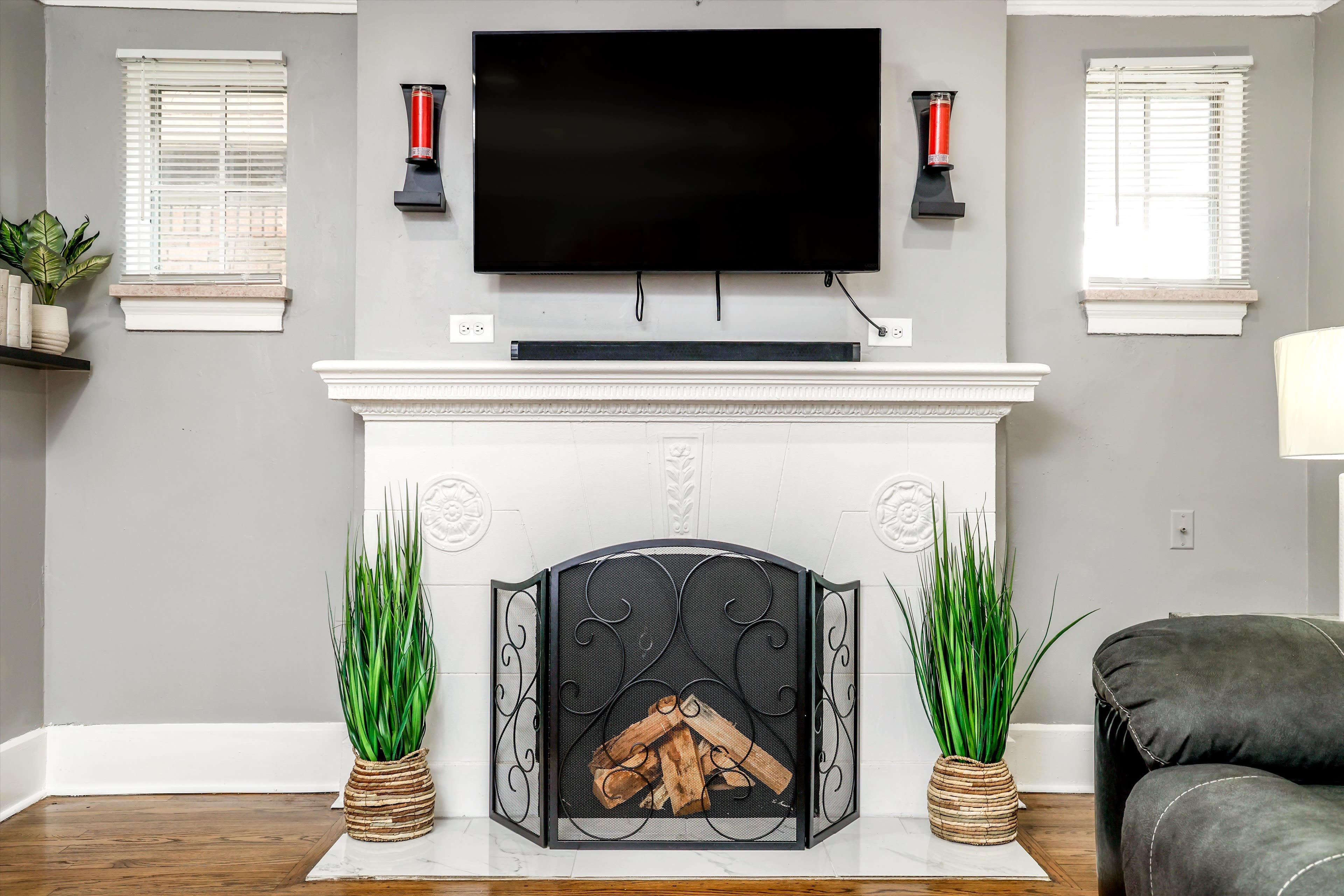 A modern living room featuring a fireplace with a mounted TV above it, flanked by tall decorative plants.
