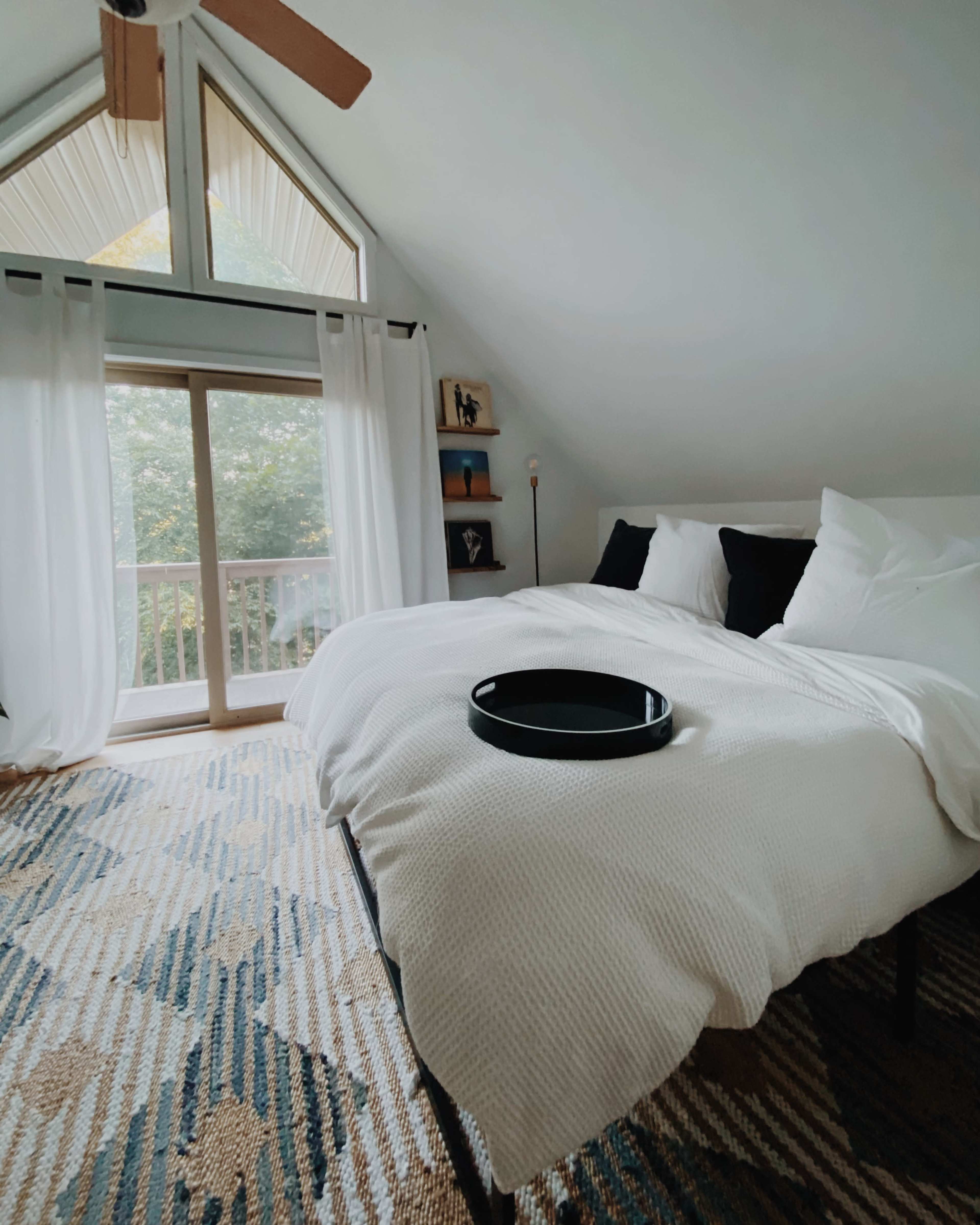 A well-lit bedroom features a bed with white linens and black accent pillows, a black tray on the bed, and large windows with sheer curtains overlooking a balcony.