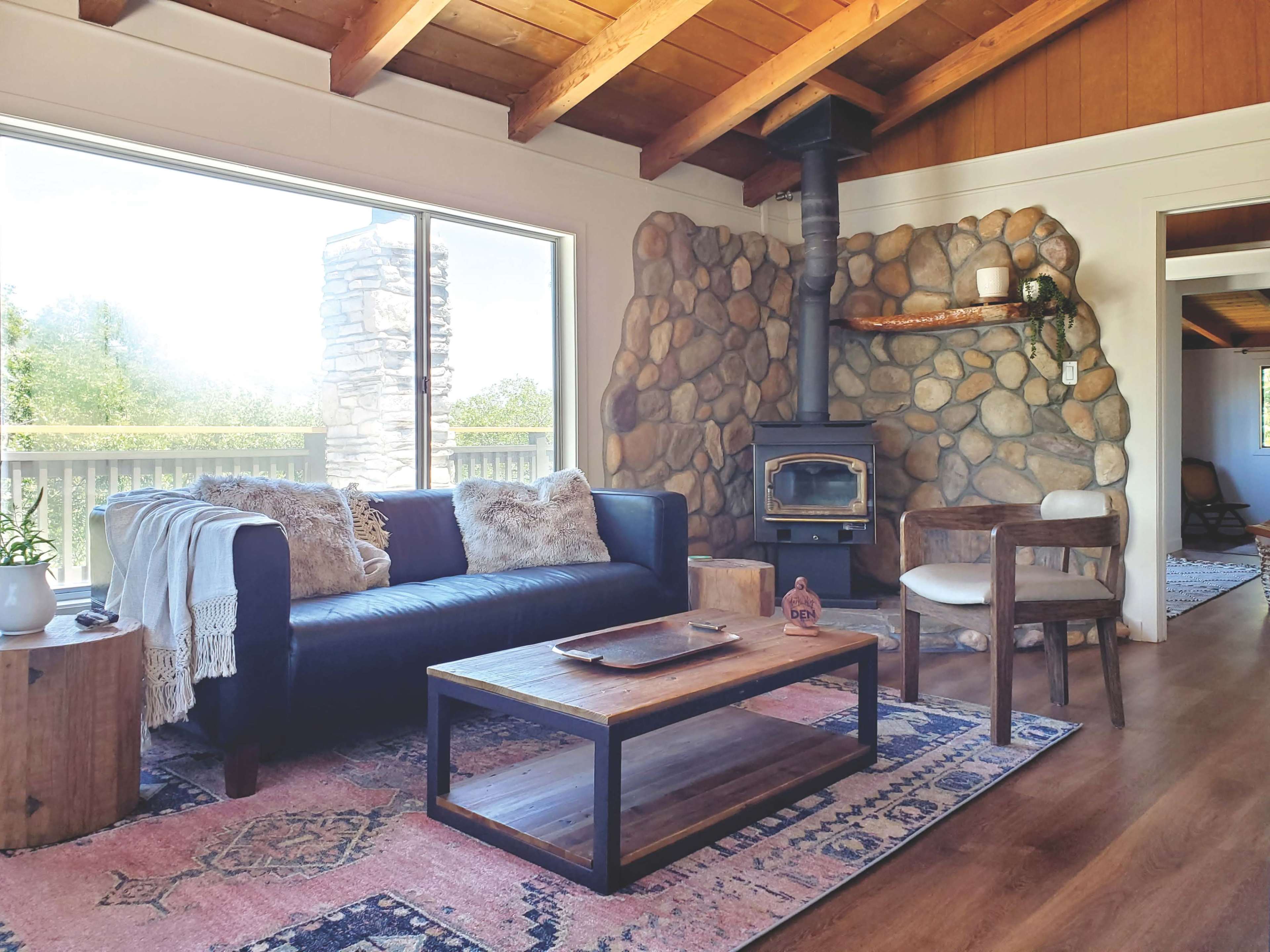 A cozy living room with a stone fireplace, a navy blue sofa, a wooden coffee table, and large windows offering natural light.