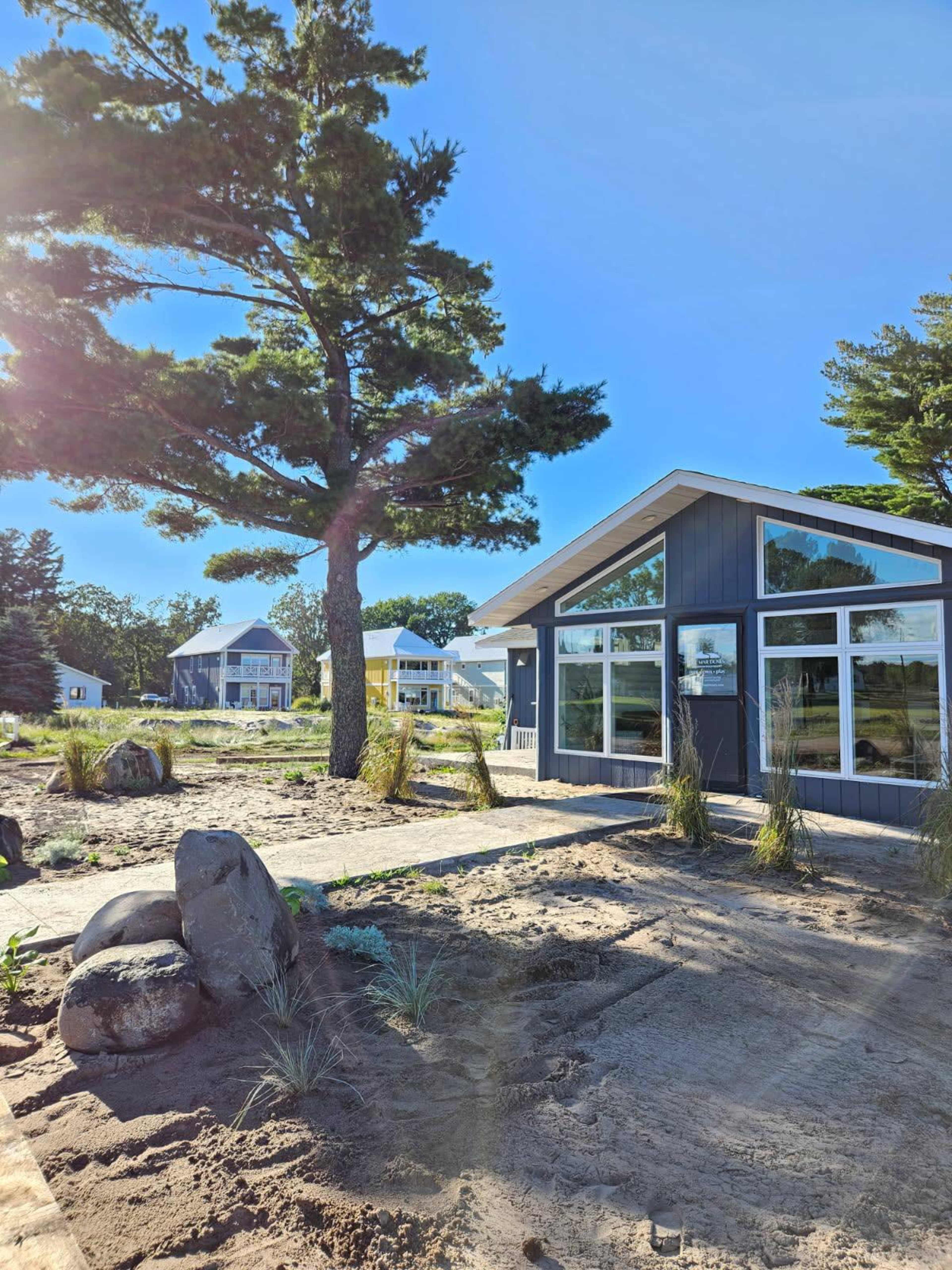 A modern gray house with large windows sits next to a sandy area, surrounded by rocks and tall grasses, under a clear blue sky.