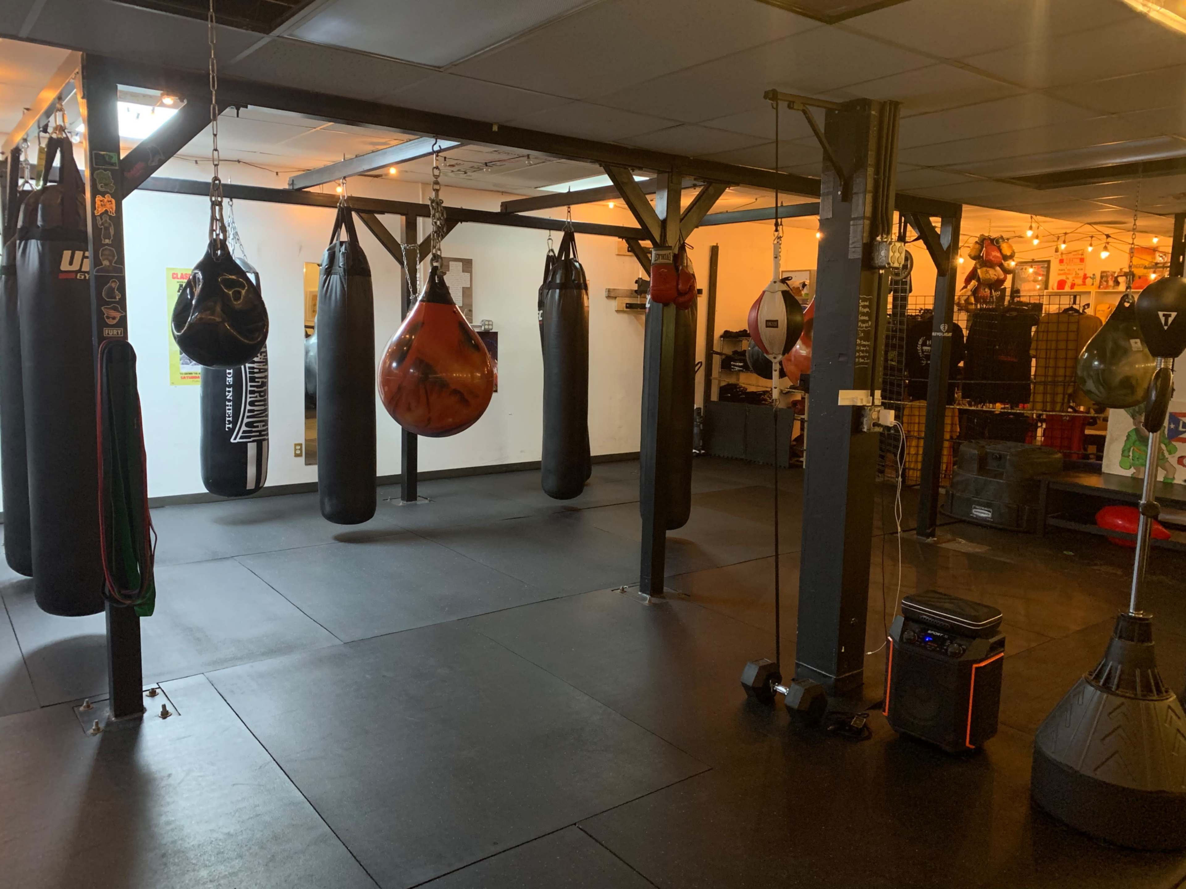 A boxing gym with several hanging punching bags, a heavy bag stand, and a black rubber flooring surface.