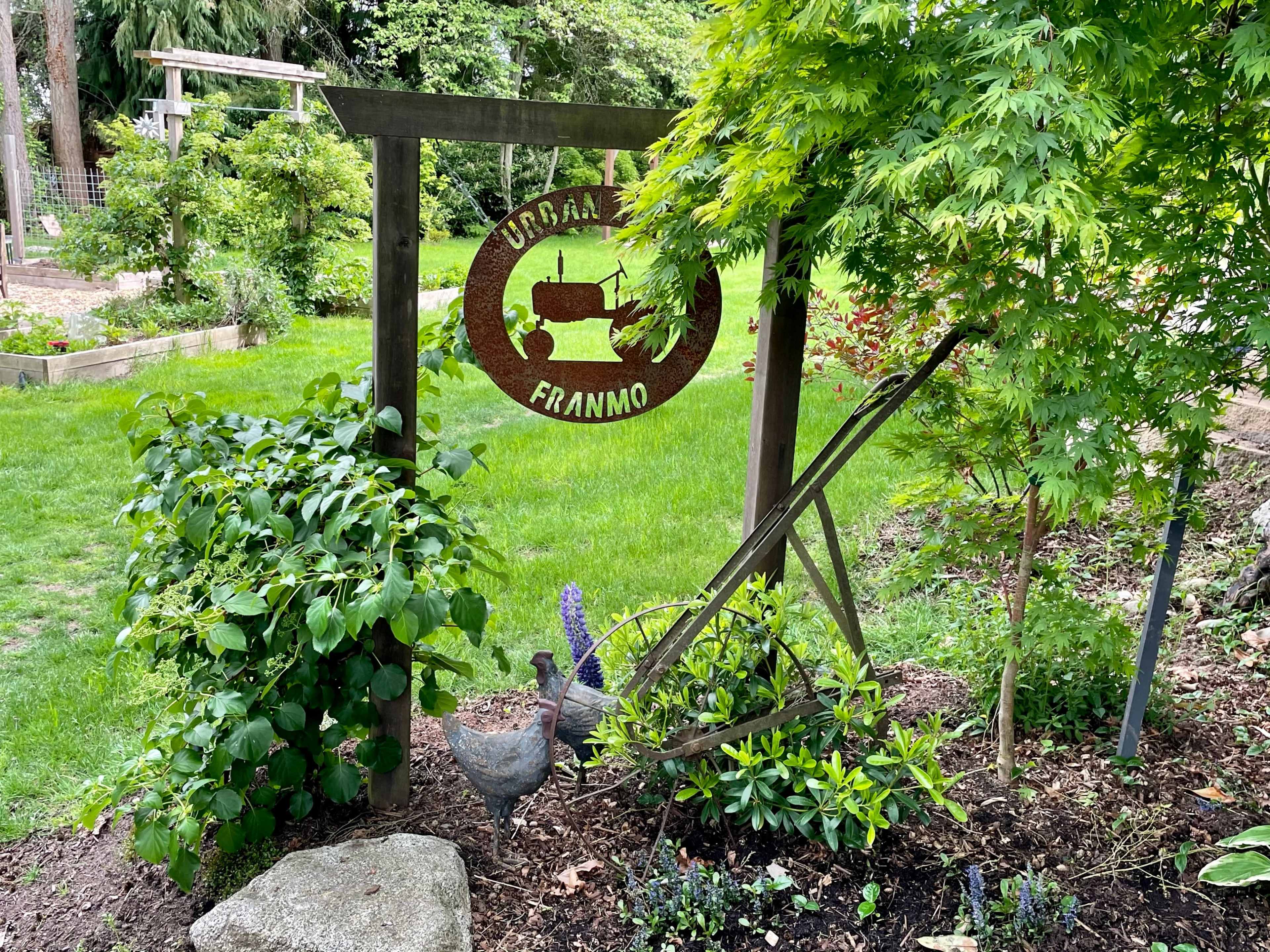 A wooden sign with "LOGAN FARM" hangs from a post amidst a garden area featuring lush greenery and a decorative chicken statue.