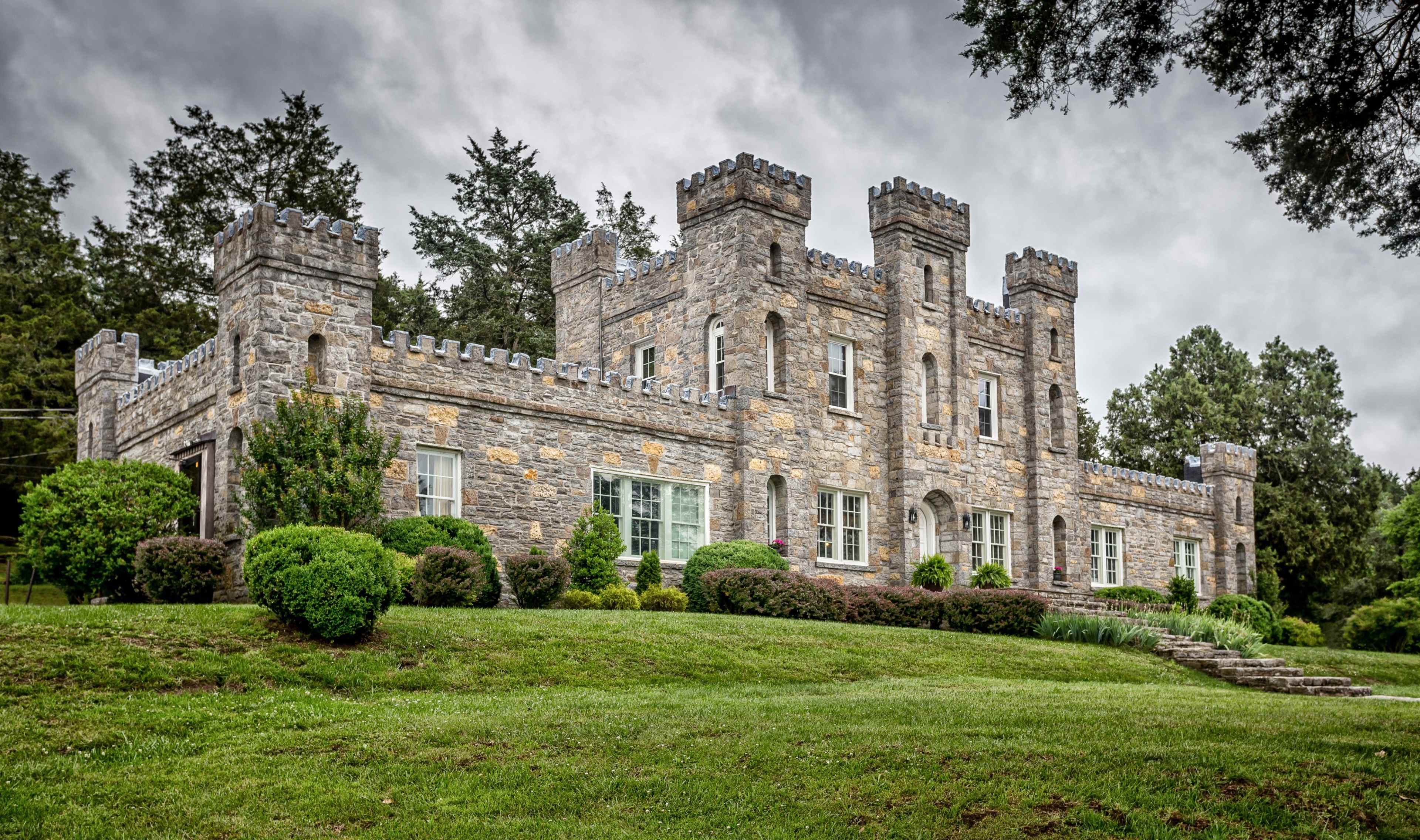A stone castle with crenelated towers and landscaped greenery is situated on a grassy hill.